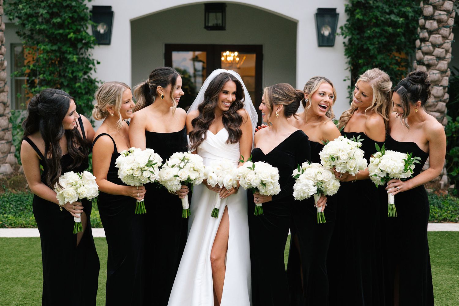 Bridal party in black dresses holding white bouquets posing outside elegant venue.