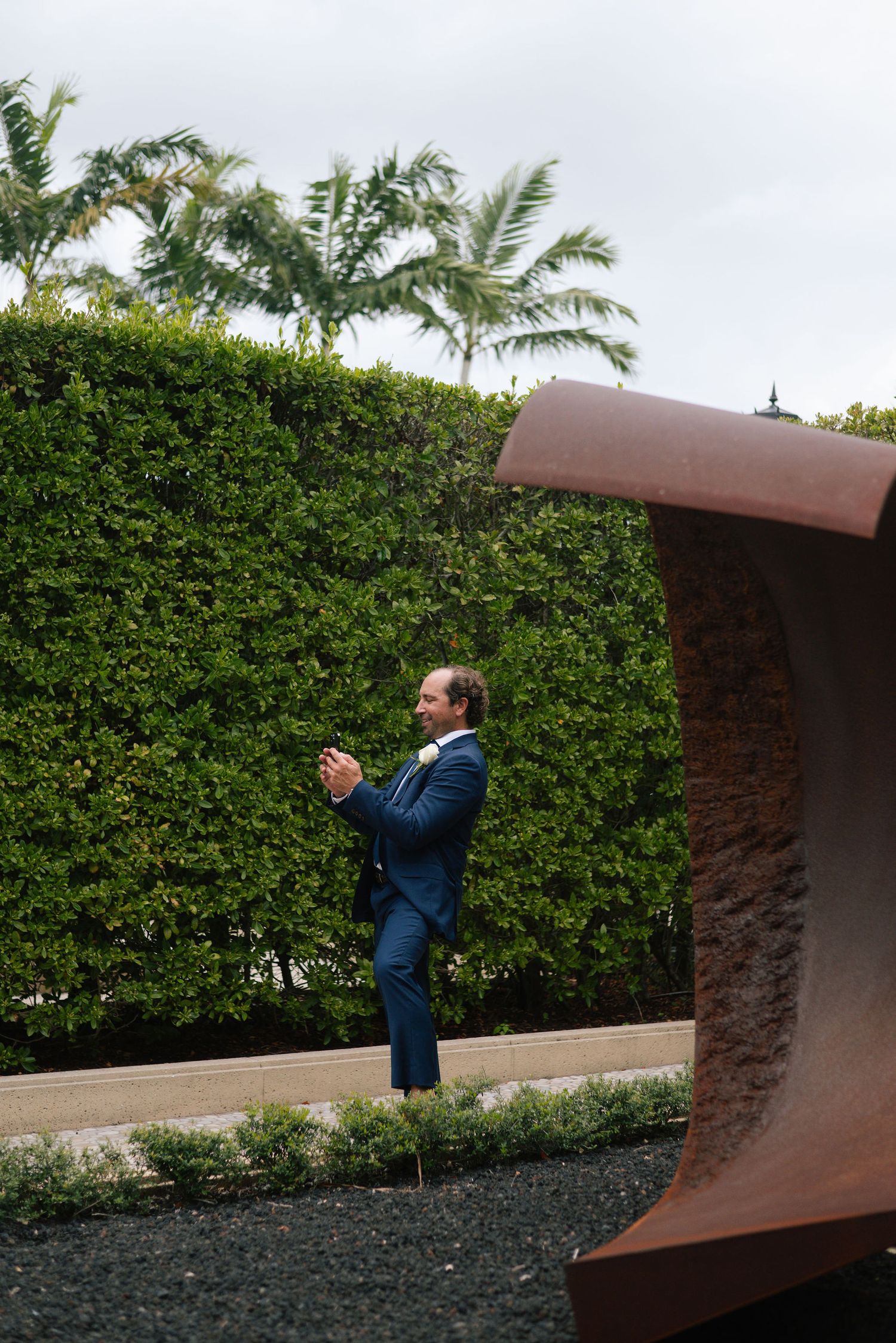 Person in navy suit walks along a curved stone wall with green hedges.