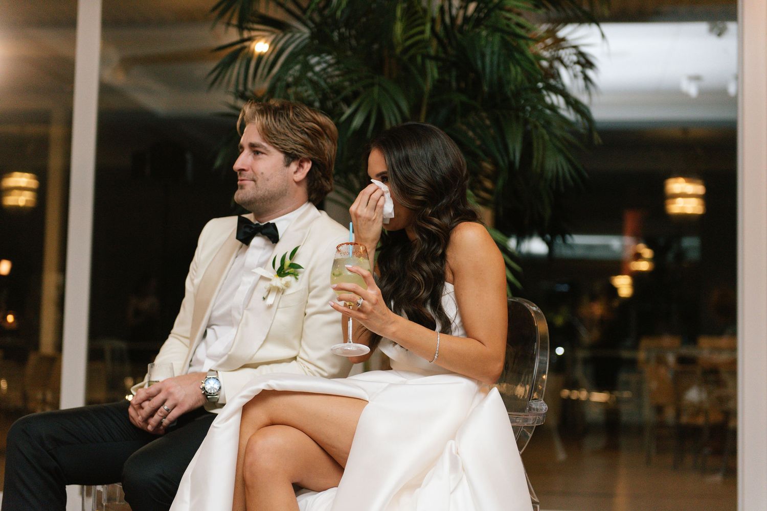 A couple embraces during their wedding reception with palm trees and ambient lighting in the background.