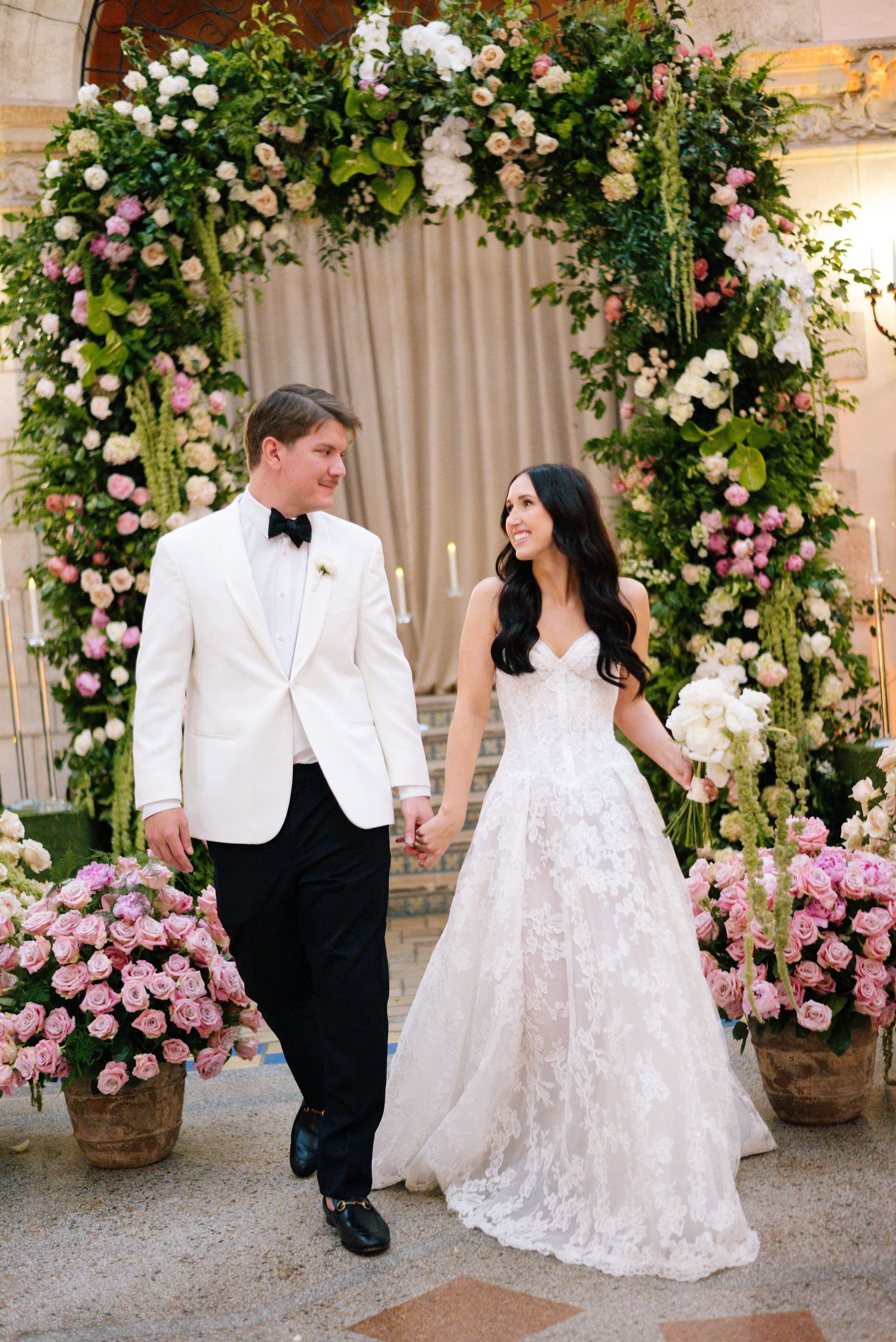 Wedding couple poses under an elegant floral arch with pink and white roses.