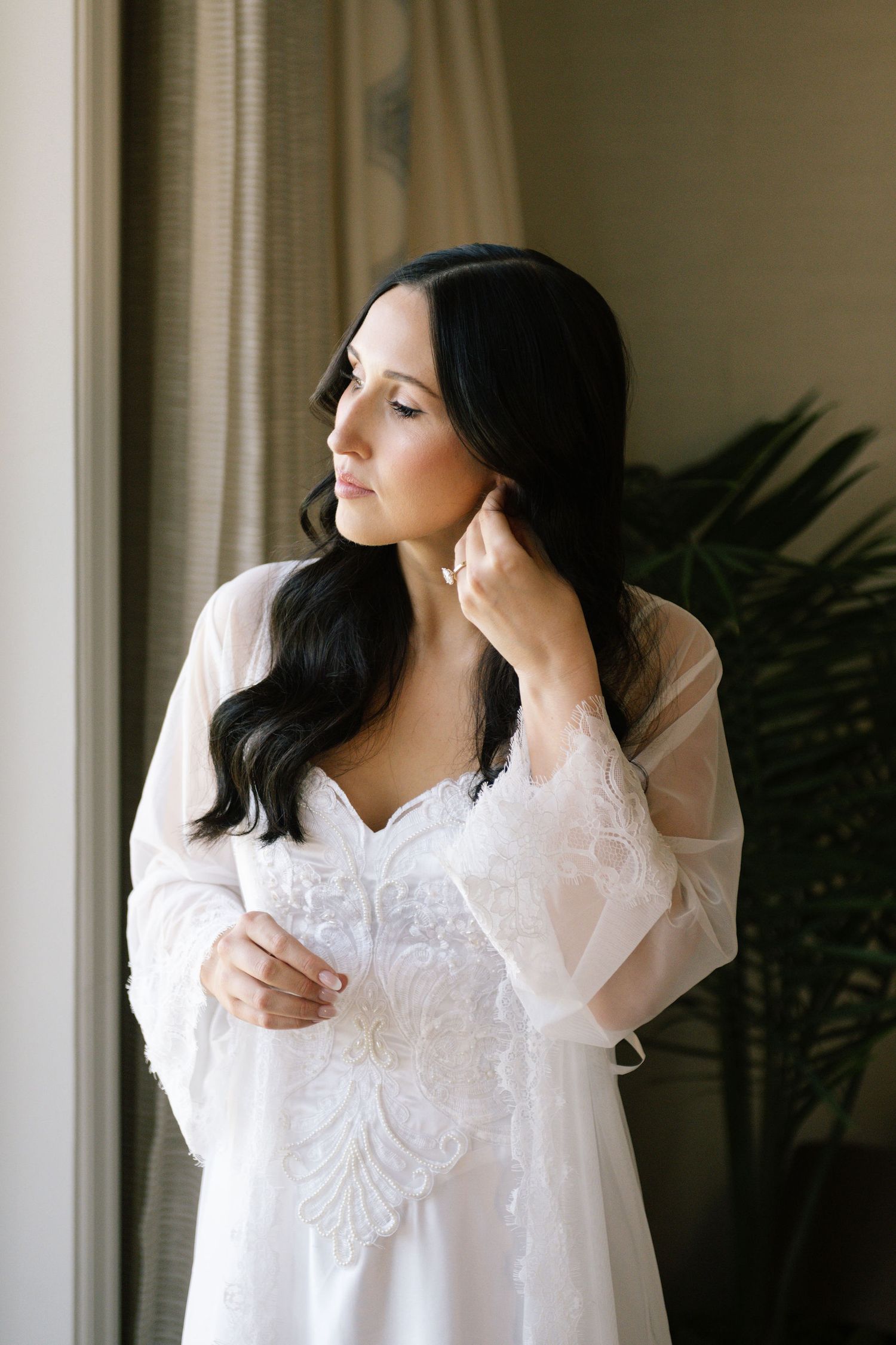 Bride in white lace robe standing by window with indoor plants.