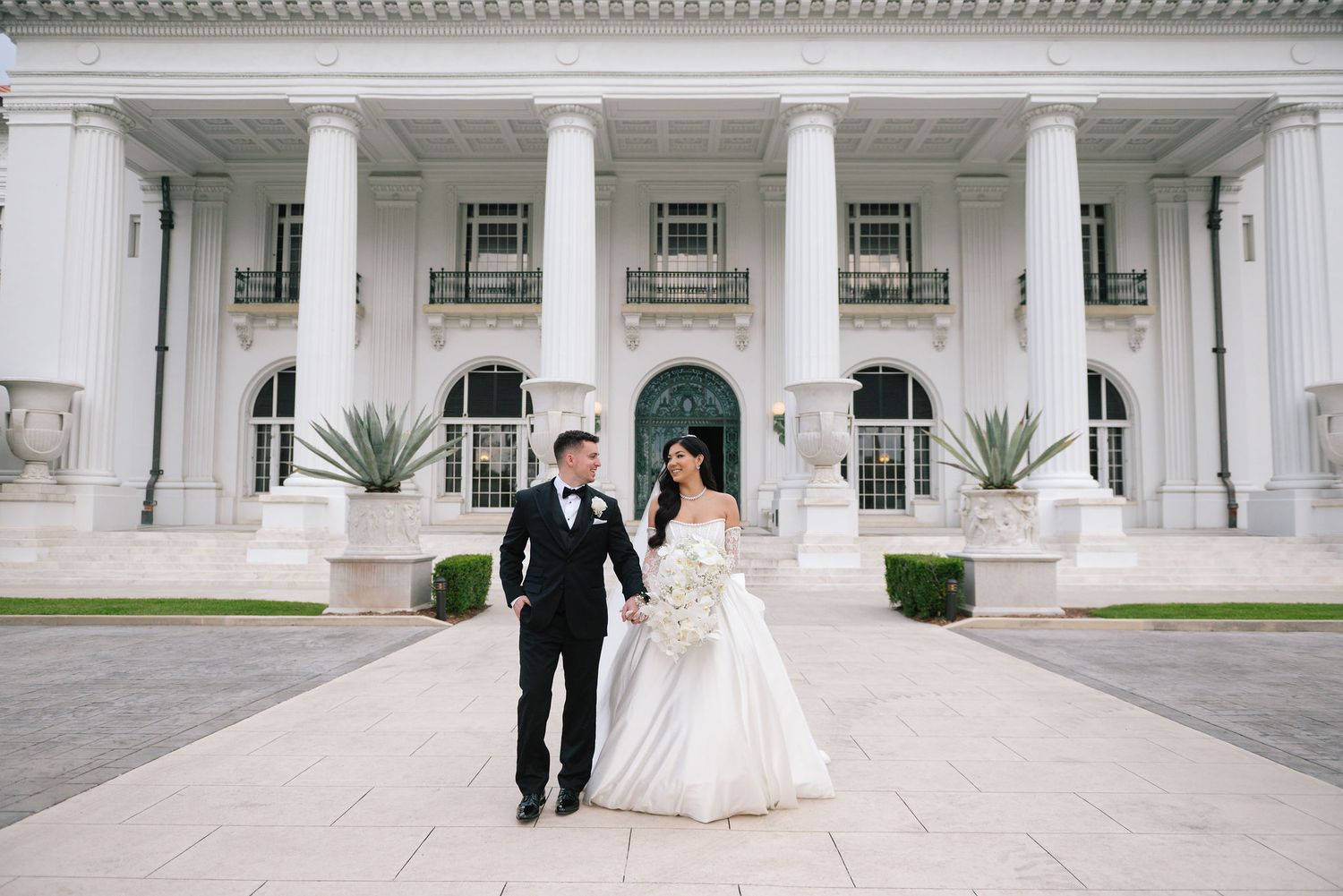 A couple walks along an elegant colonnade with white pillars at a grand venue.