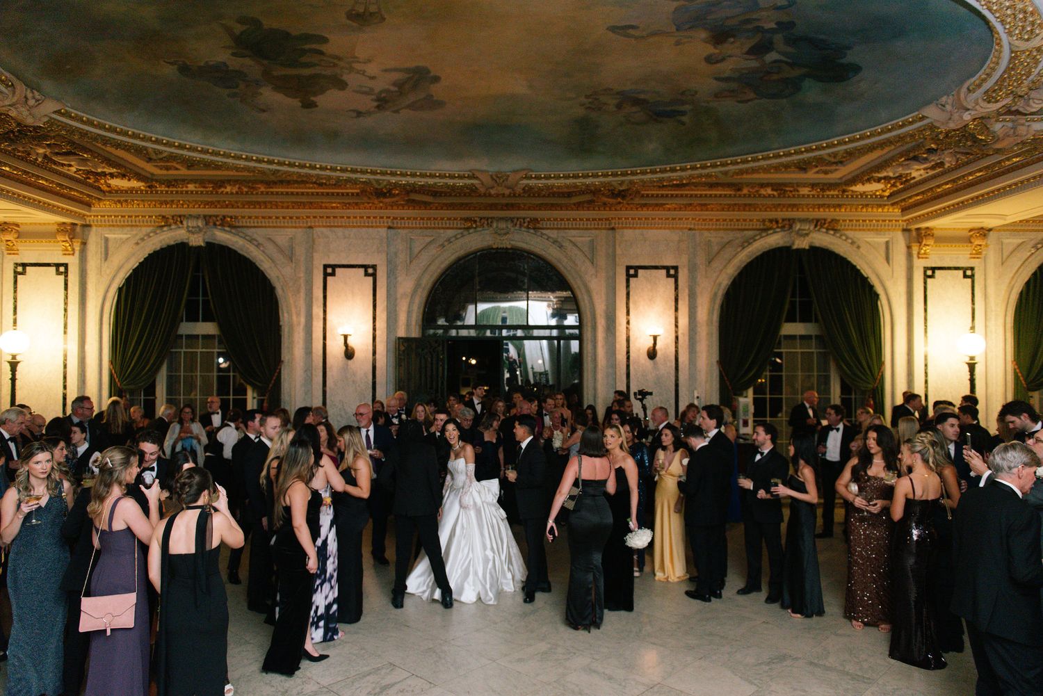 Grand ballroom wedding reception with ornate ceiling and crowd of formal guests.