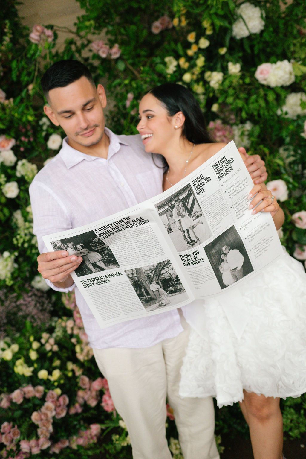 Couple in white attire sharing a newspaper against a rose-covered wall.