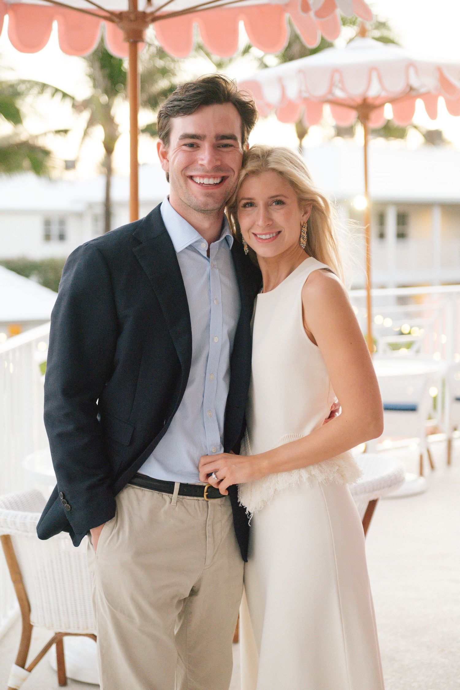Elegant couple dressed in formal attire pose together on a balcony with pink and white striped umbrellas overhead.