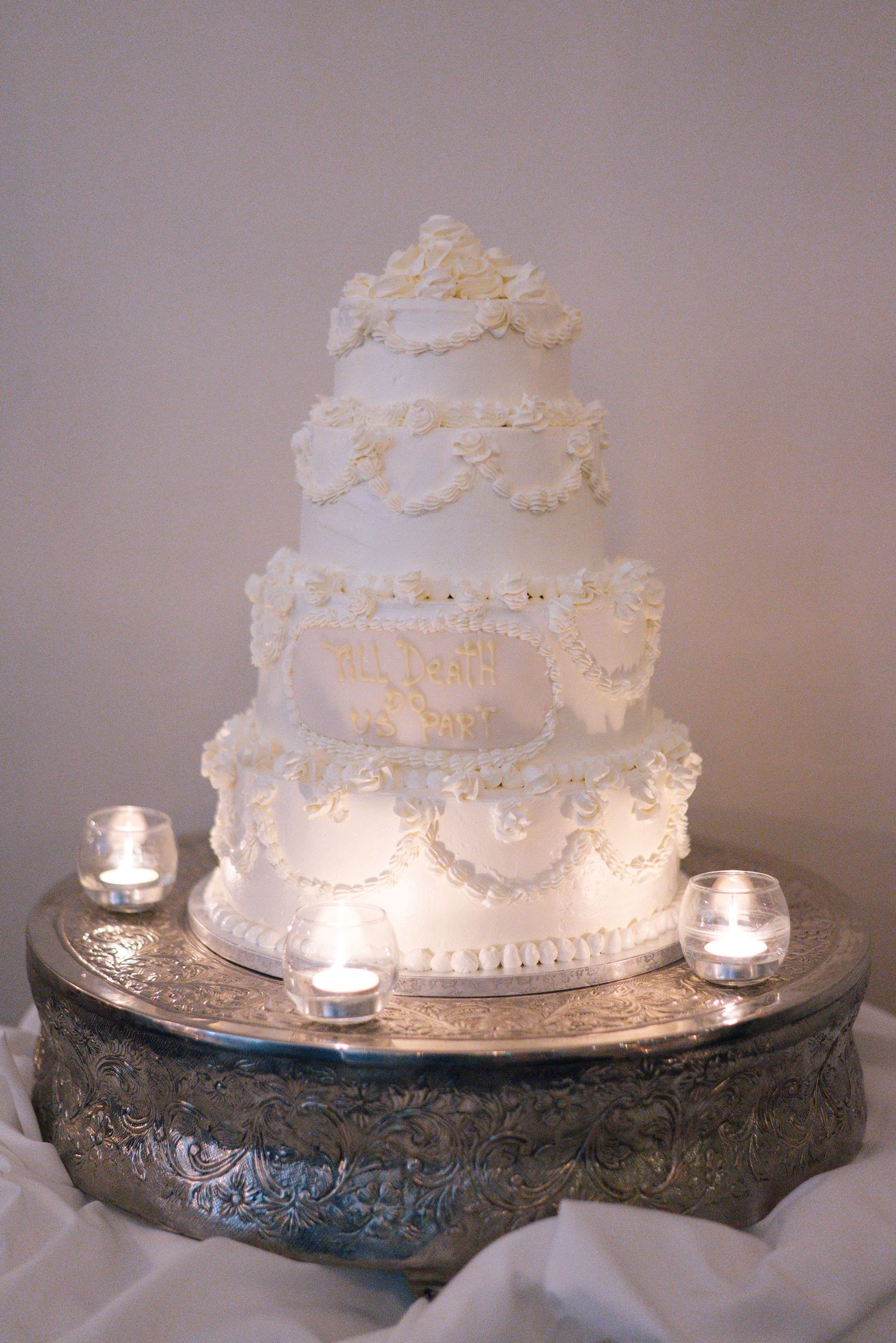 Elegant white wedding cake with intricate piping details illuminated by soft candlelight on an ornate silver stand.