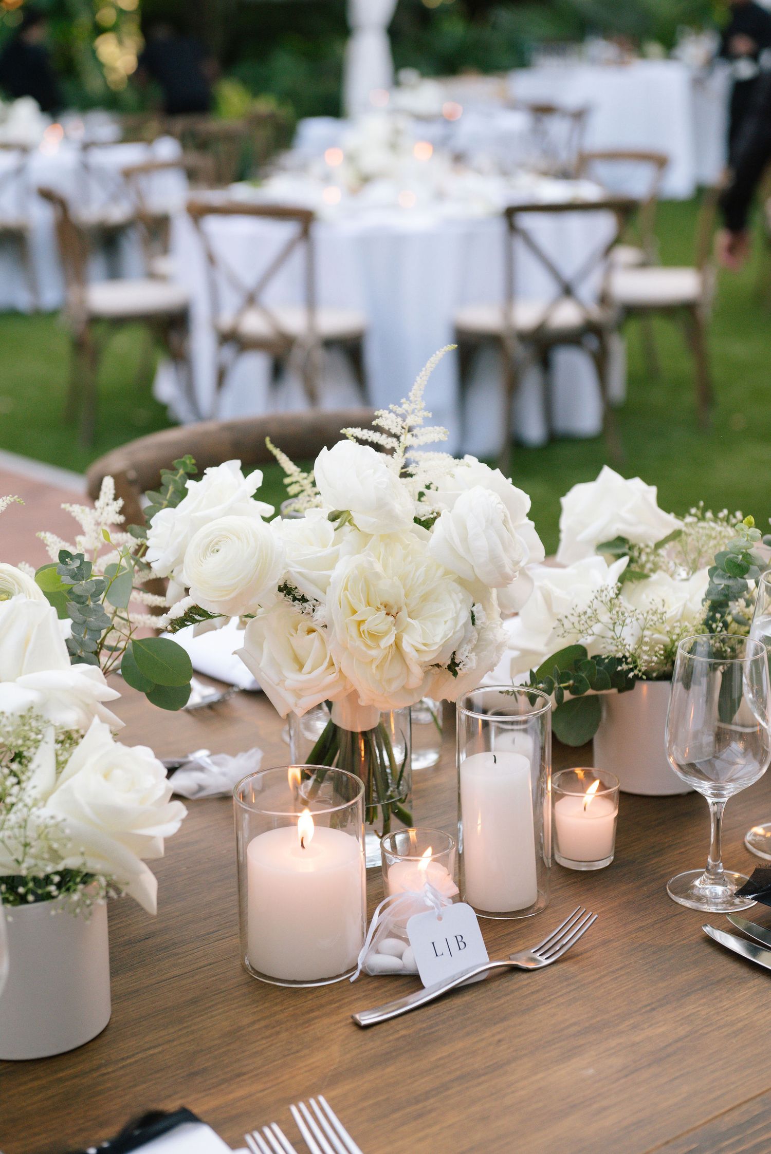 Elegant wedding table setting with white floral centerpieces, glowing candles and rustic wooden chairs in an outdoor garden.