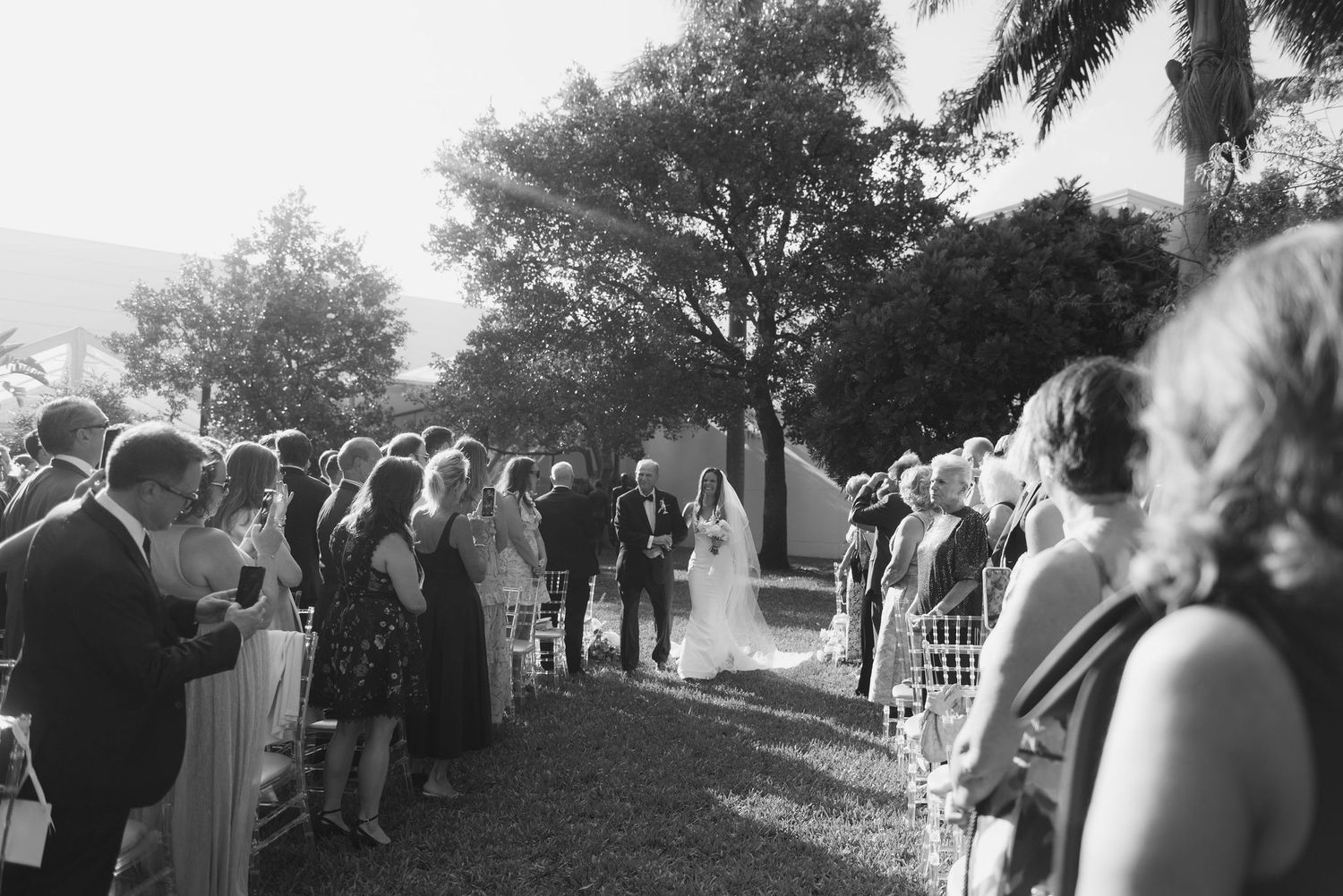 Wedding guests line both sides of an outdoor aisle beneath trees during a daytime ceremony.
