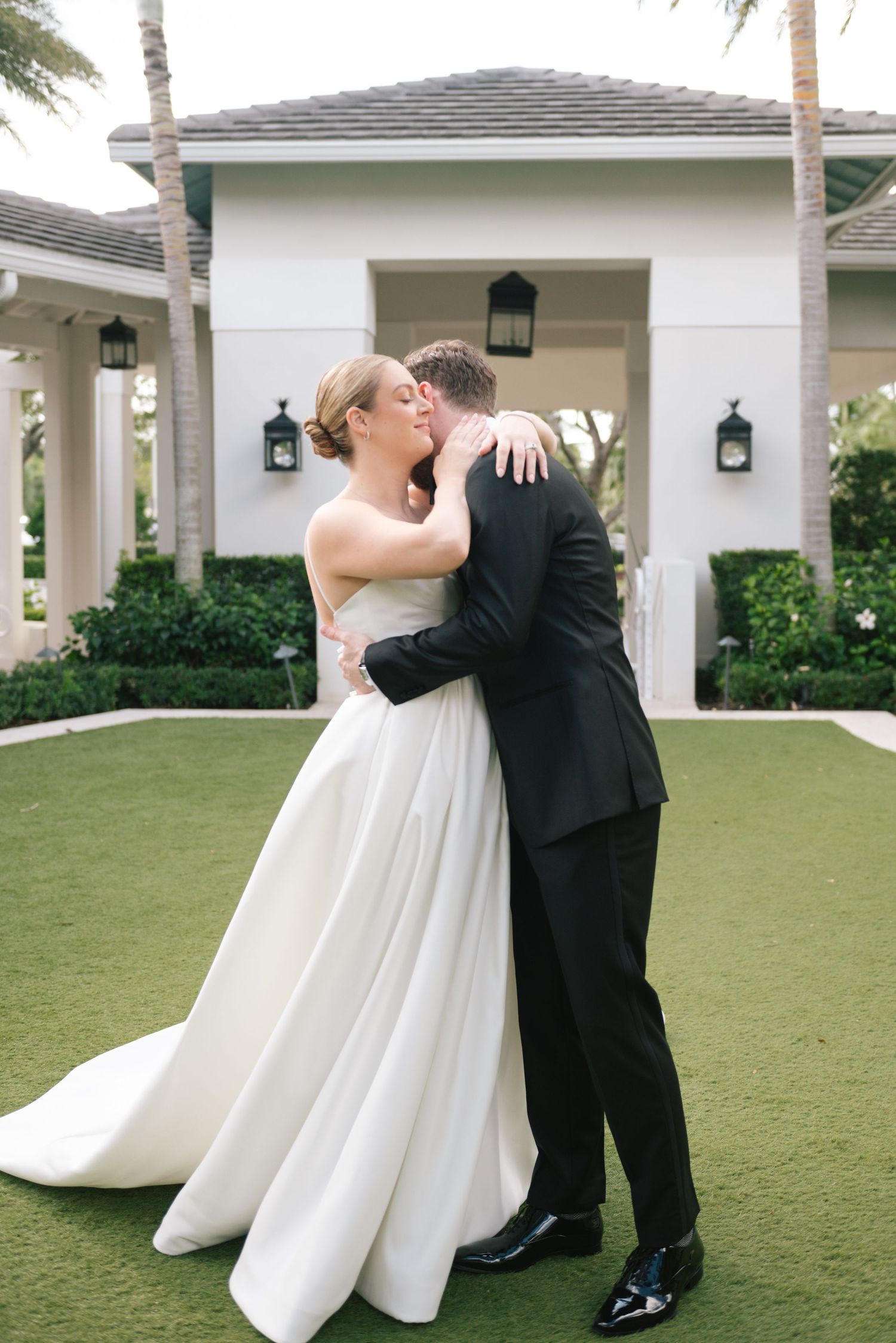 A romantic wedding couple embraces and dances on a lush green lawn with palm trees in the background.