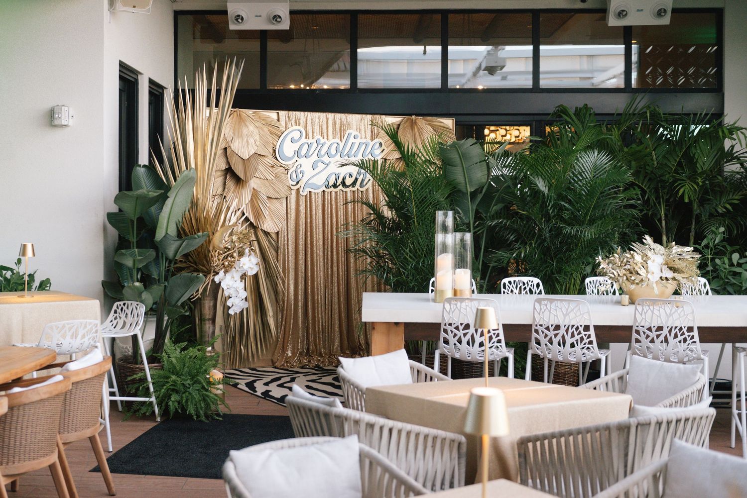 Modern coastal restaurant interior with white woven chairs, natural wood tables and lush tropical plants.