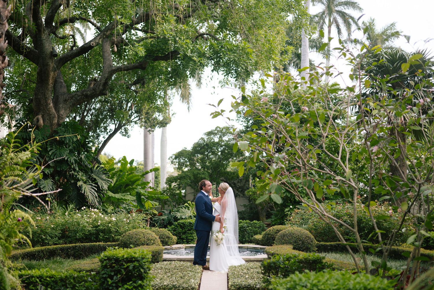 Couple sharing a romantic moment in a lush tropical garden with tall palm trees and manicured landscaping.
