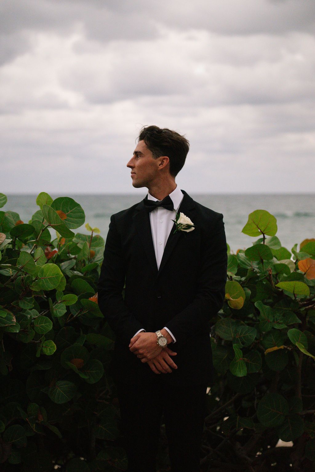 Groom in classic black tuxedo poses against coastal landscape.