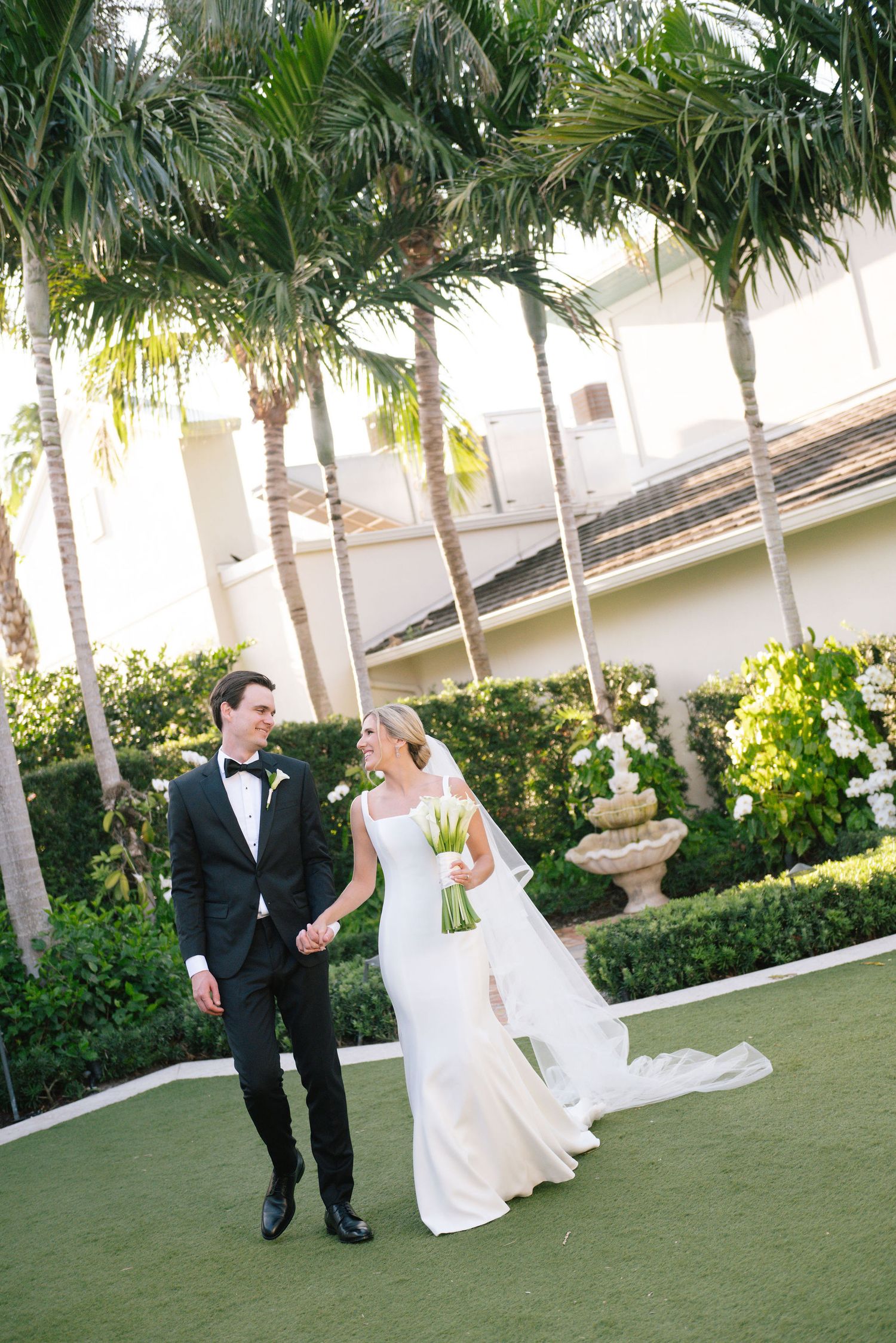 Wedding couple walking together on lawn lined with tall palm trees.