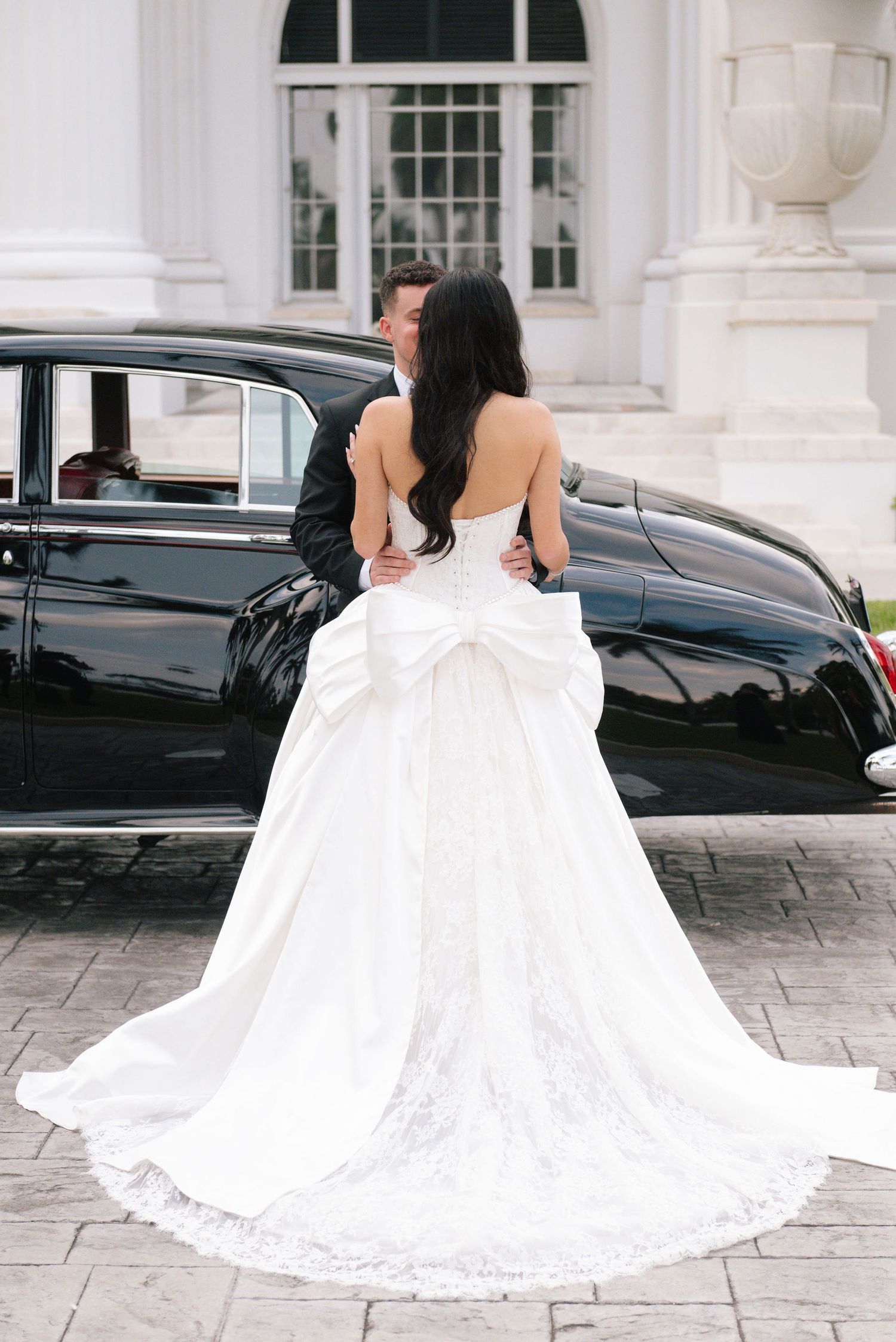 A flowing white wedding dress with intricate lace details photographed from behind on stone pavement.