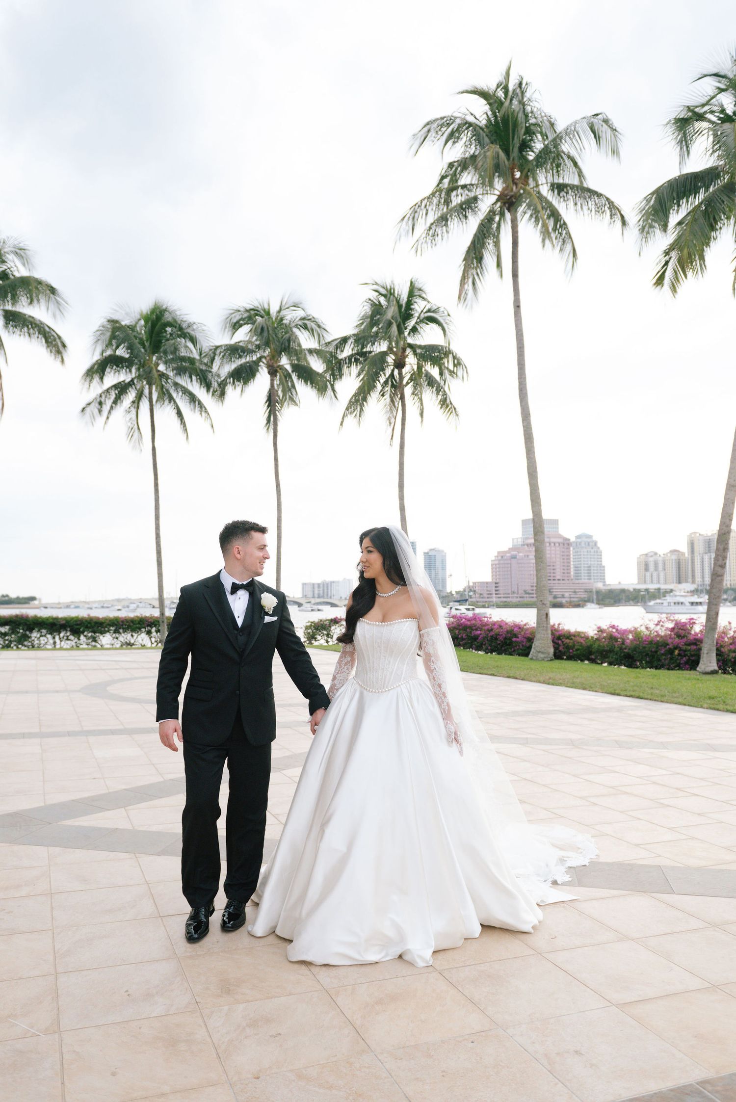 A wedding couple walks hand in hand along a palm-lined pathway with cityscape views.