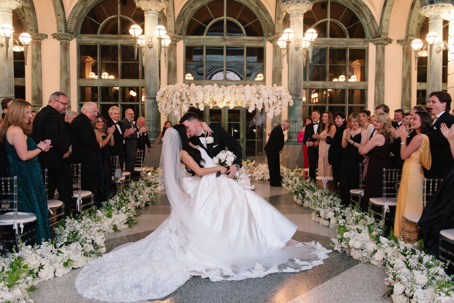 Elegant wedding ceremony with white floral arrangements and guests seated in grand ballroom.