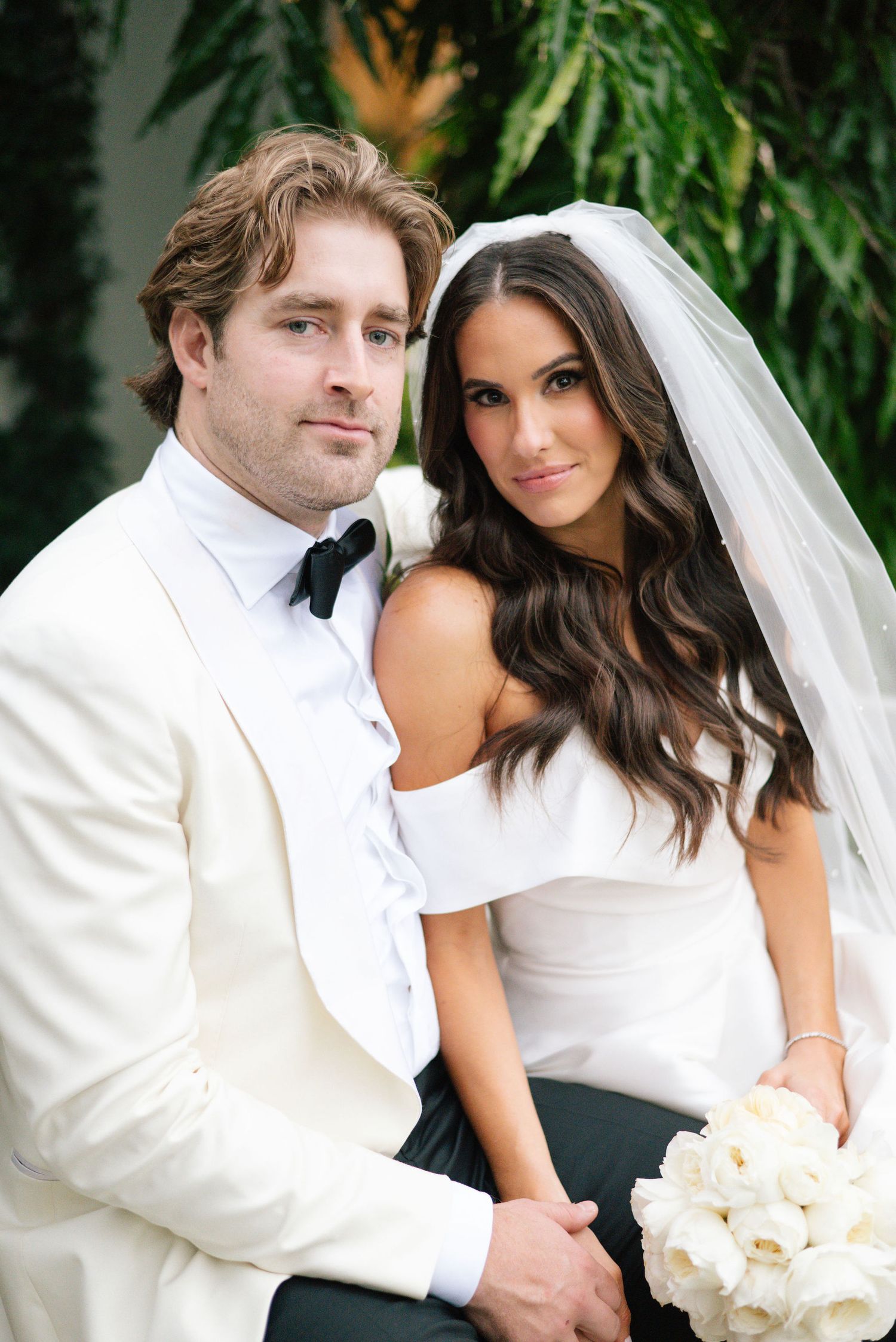 Elegant wedding couple poses together in white attire with a bridal bouquet against a lush green backdrop.