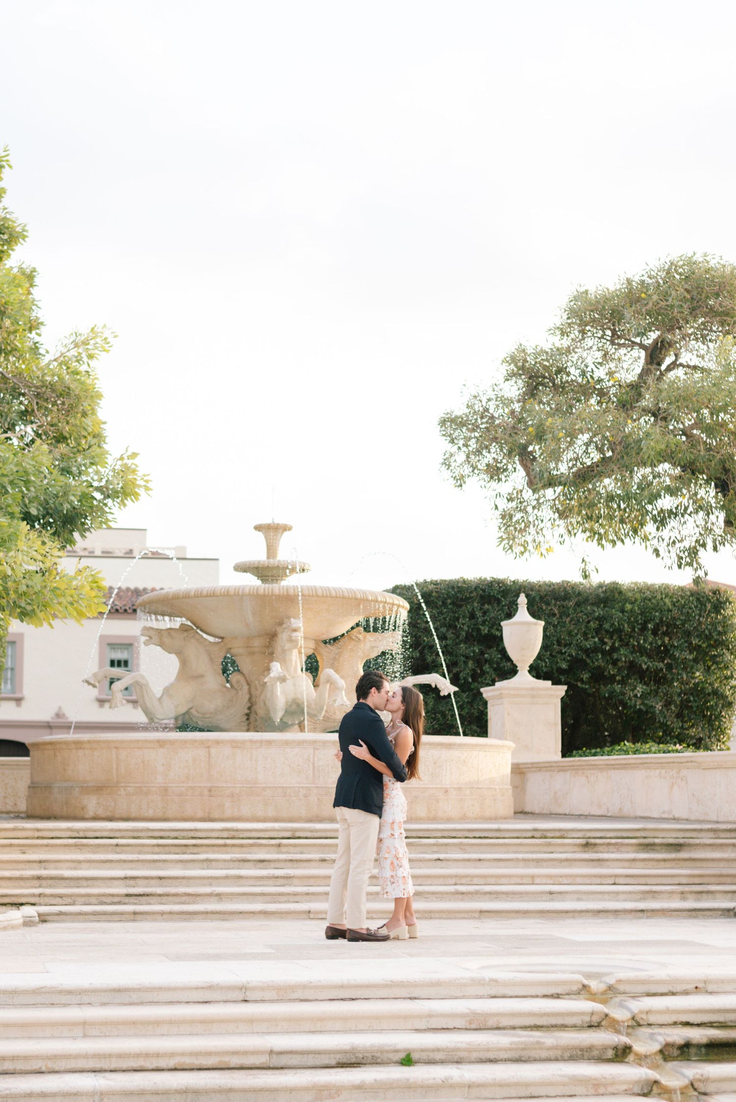 A couple embraces on elegant stone steps near a decorative fountain and manicured hedges in a formal garden setting.