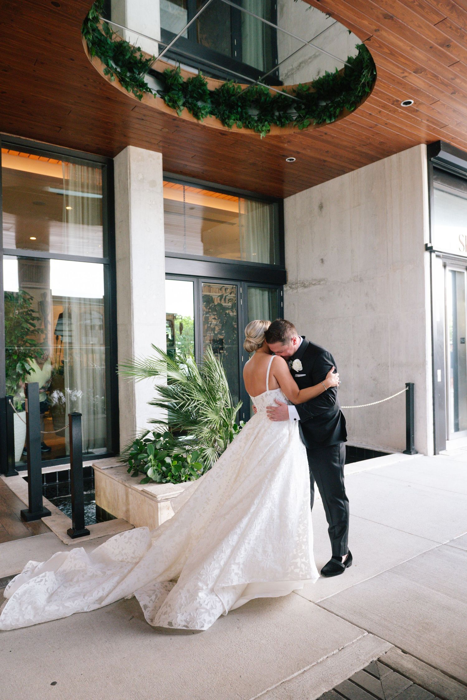 A bride and groom share a romantic kiss outside a modern wedding venue adorned with lush greenery.