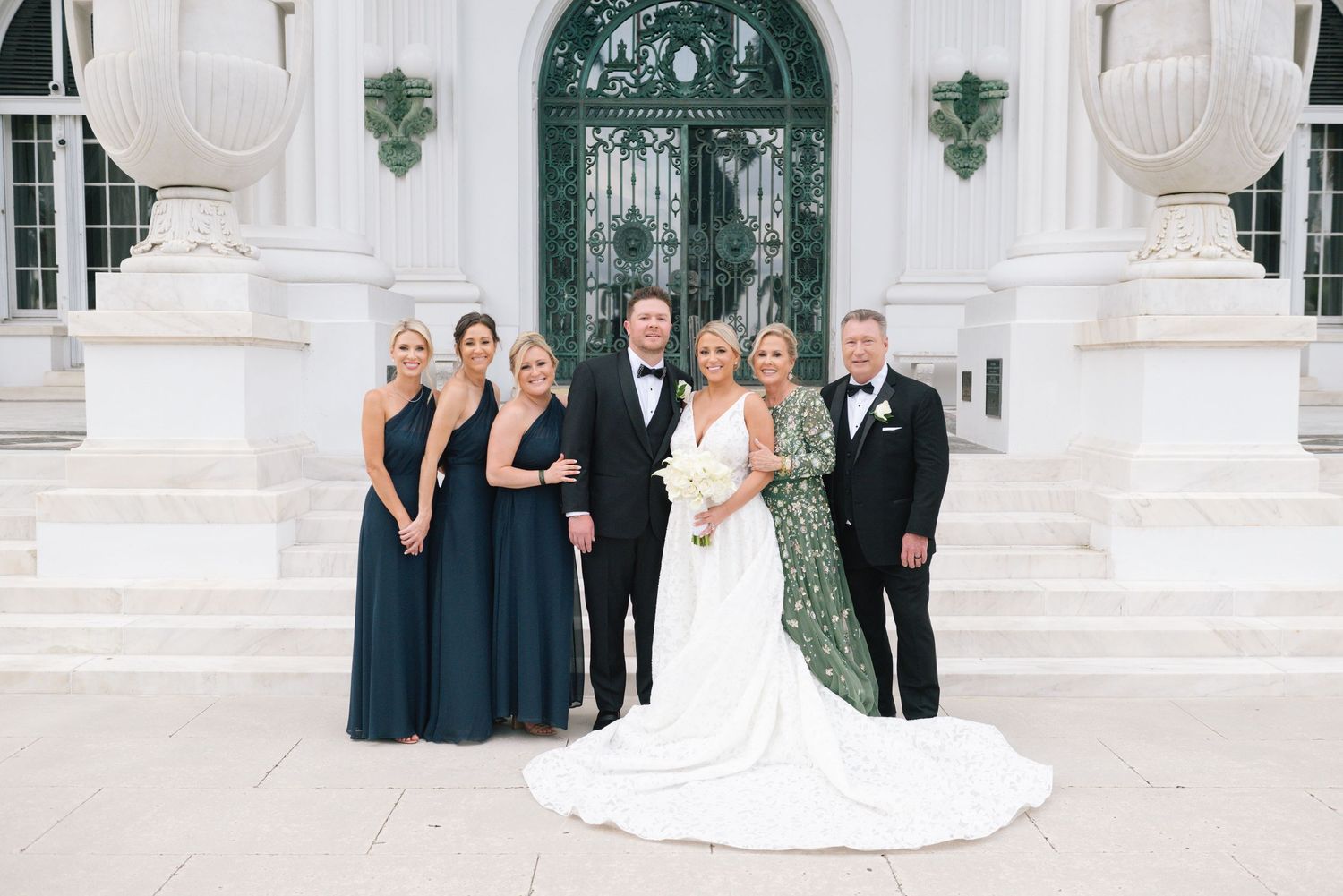 Wedding party poses in navy gowns and tuxedos before elegant white columns and ornate green doors.
