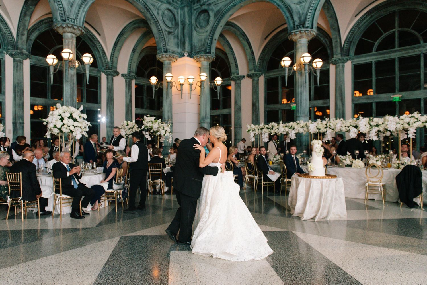 Bride and groom share first dance in ornate ballroom with white floral centerpieces.