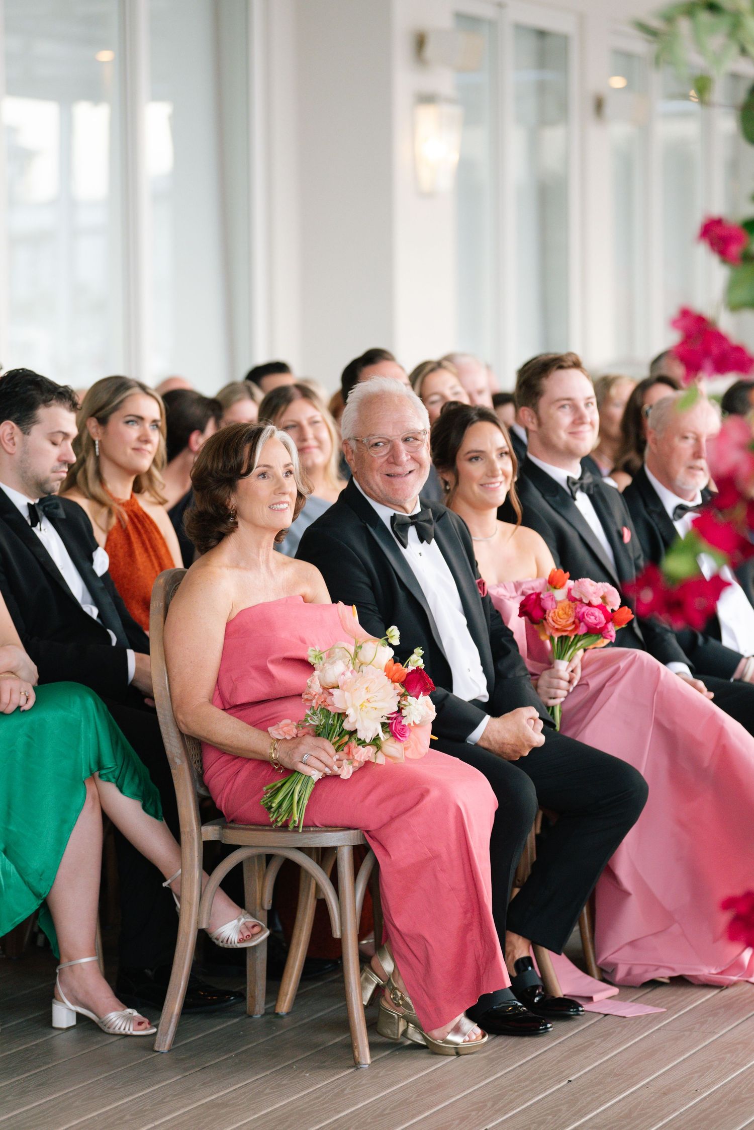Wedding guests seated in formal attire with pink and green dresses in bright indoor venue.