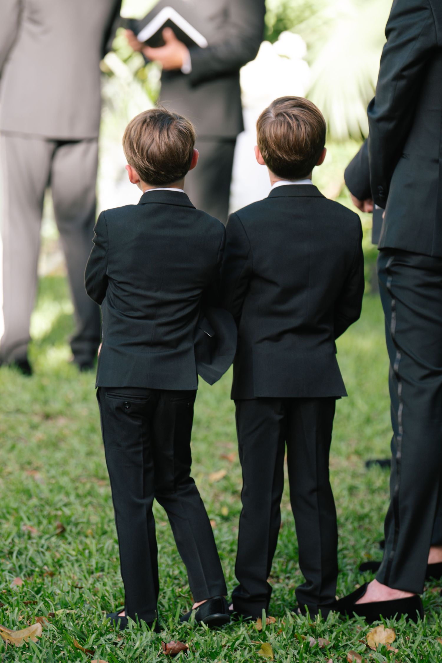 Young boys in black suits standing on grass at a formal outdoor event.