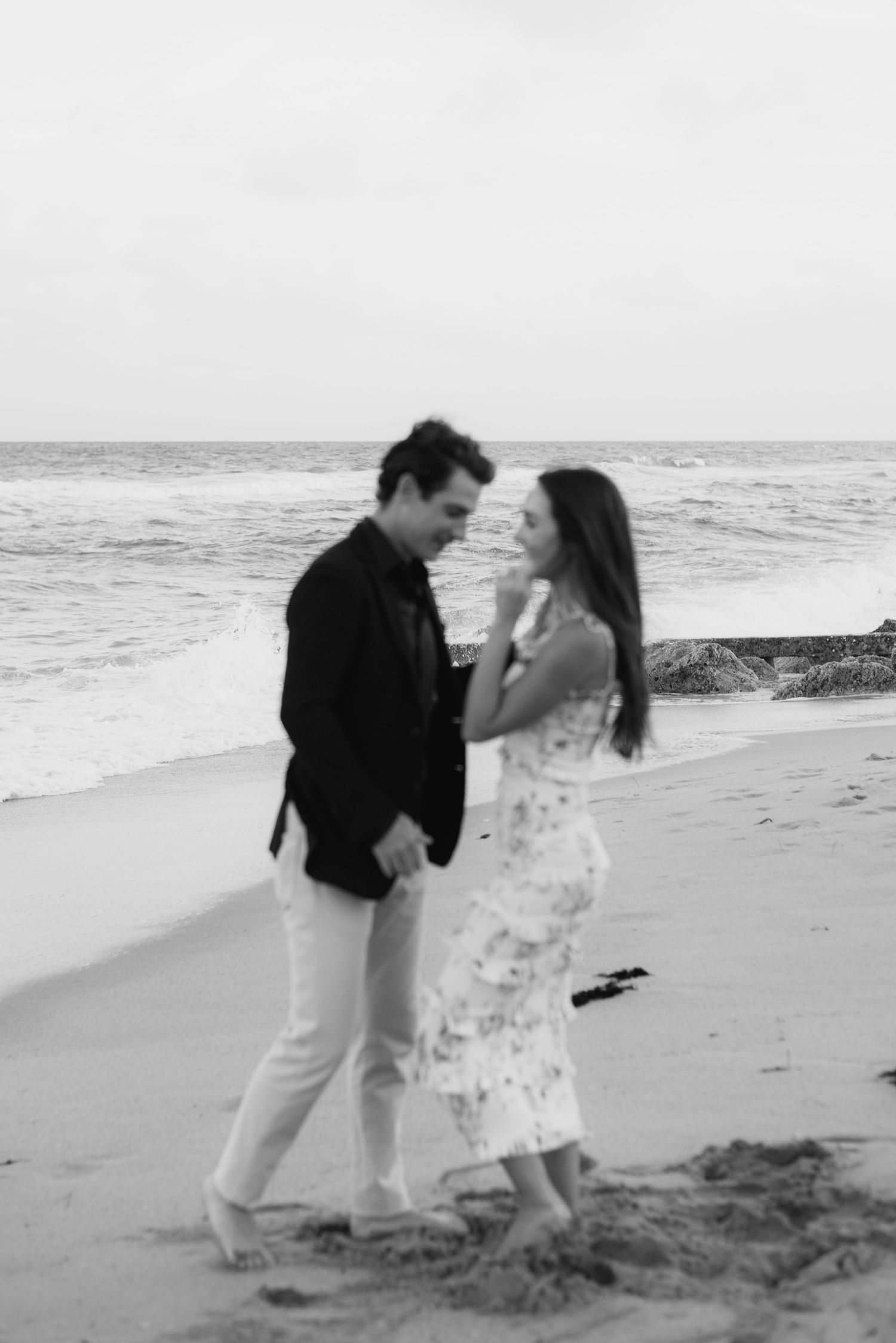 A couple walks along a sandy beach at sunset in black and white, creating a romantic silhouette against the ocean.