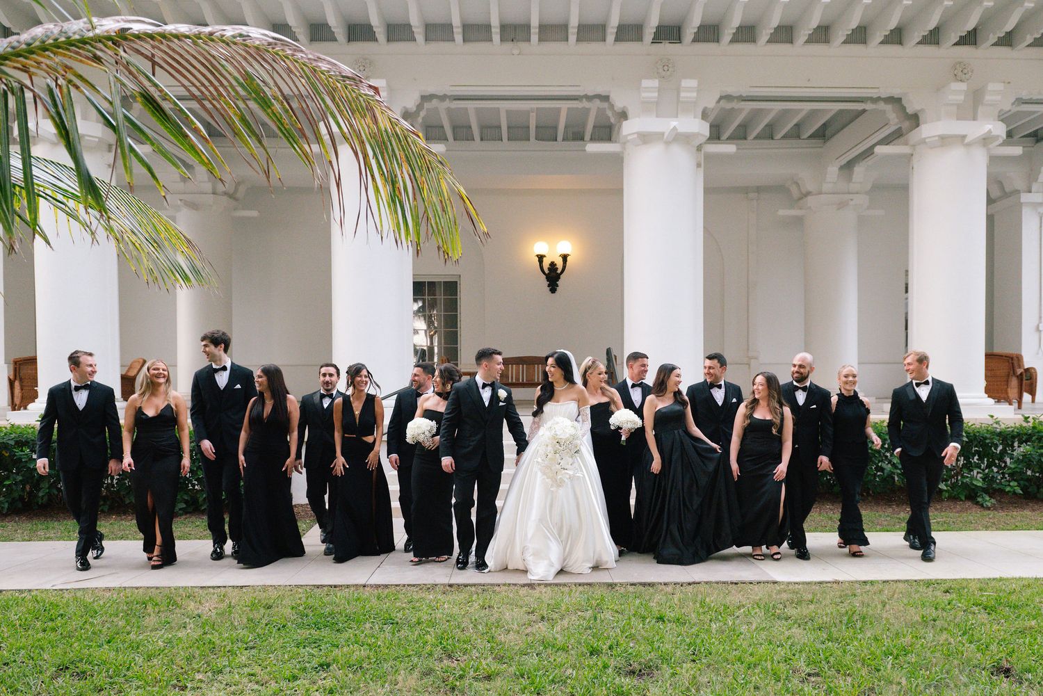 Wedding party poses under white columns with palm trees in the background.