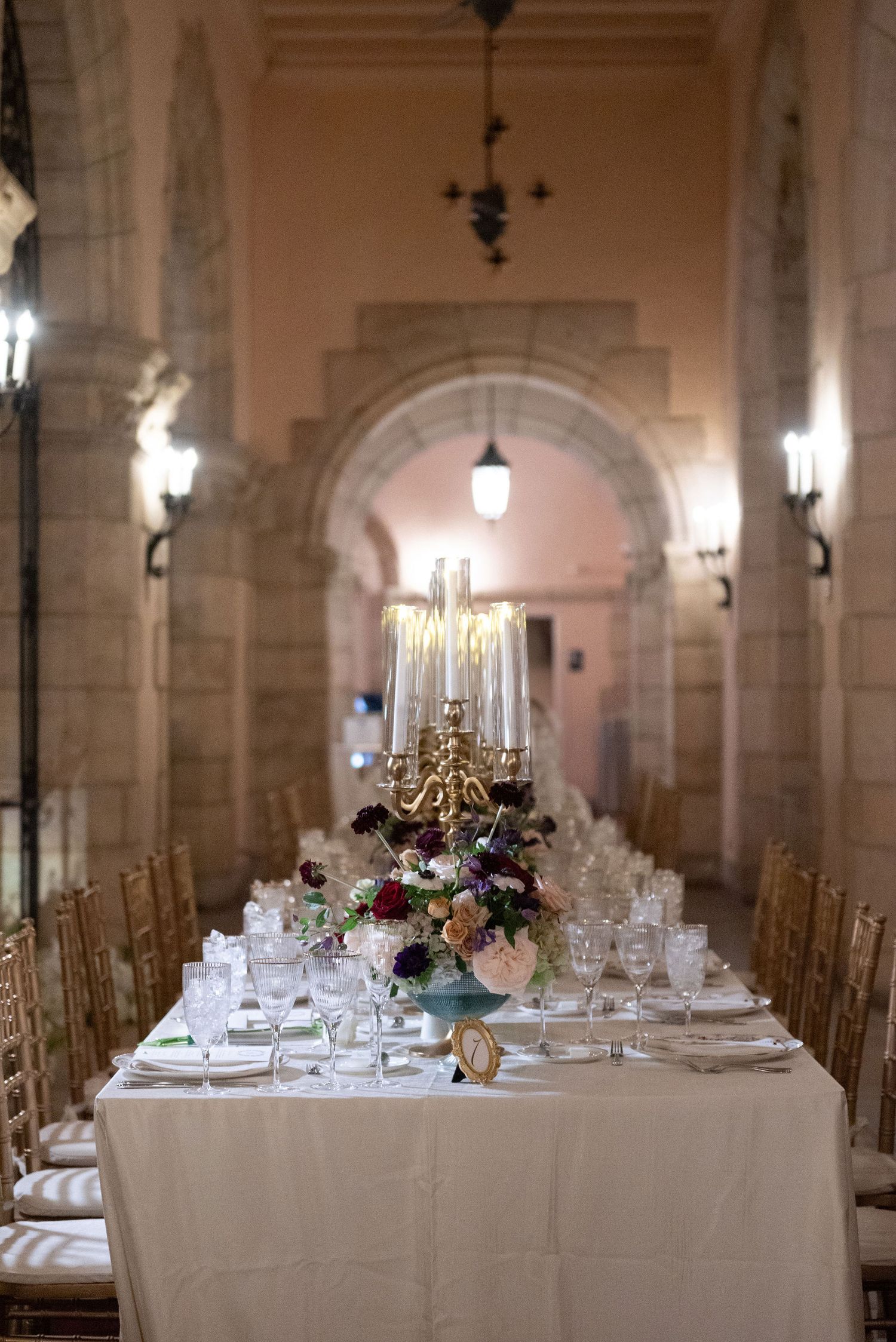 Long banquet tables set for a formal wedding reception in a grand stone-walled venue with arched architecture.