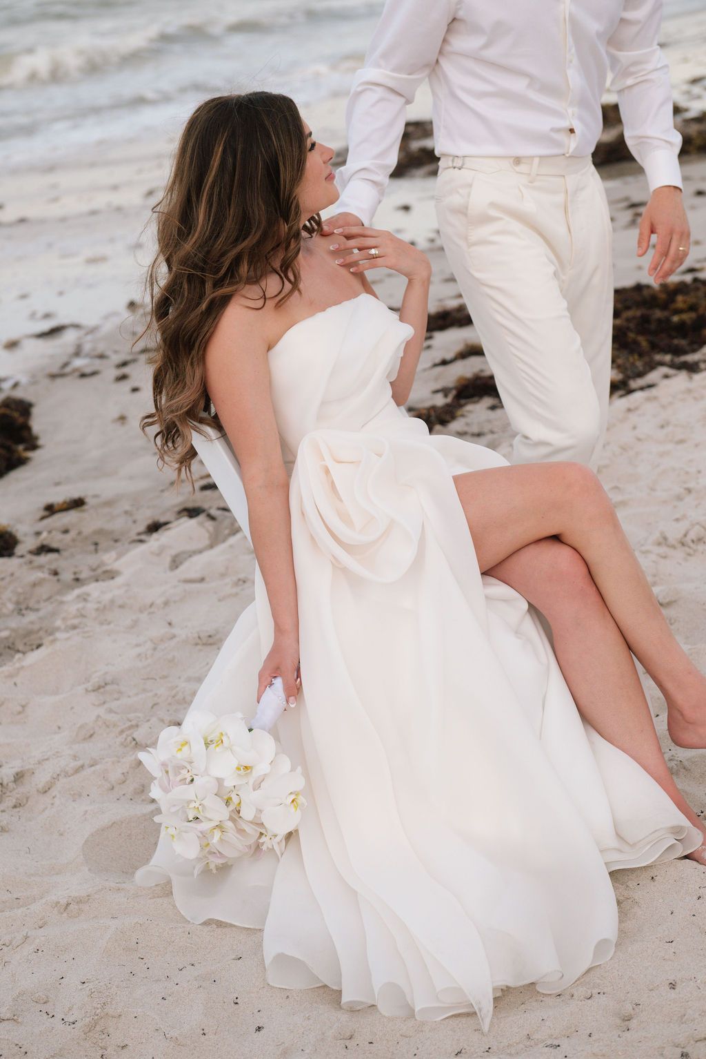 A bride in a flowing white dress sits on a sandy beach holding a bouquet of white orchids.