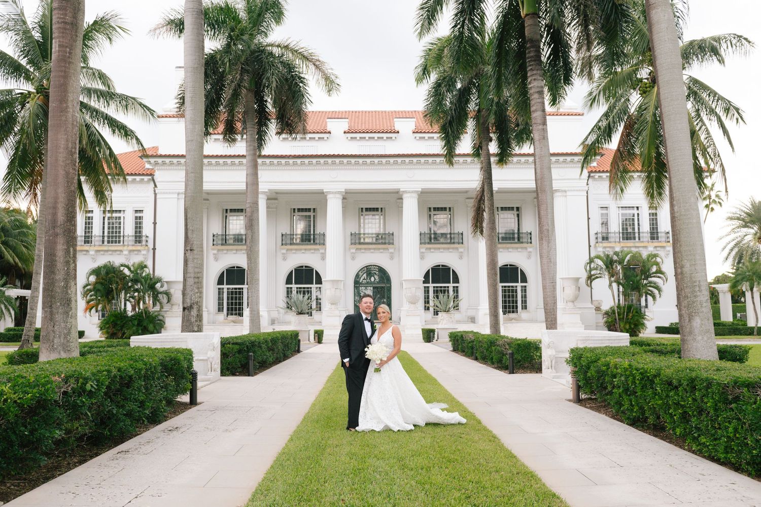 Bride and groom pose in front of a grand white mansion surrounded by tall palm trees and manicured hedges.