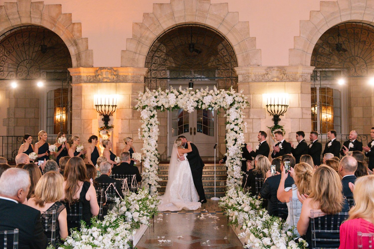 Bride and groom kiss under white floral arch during romantic indoor wedding ceremony.