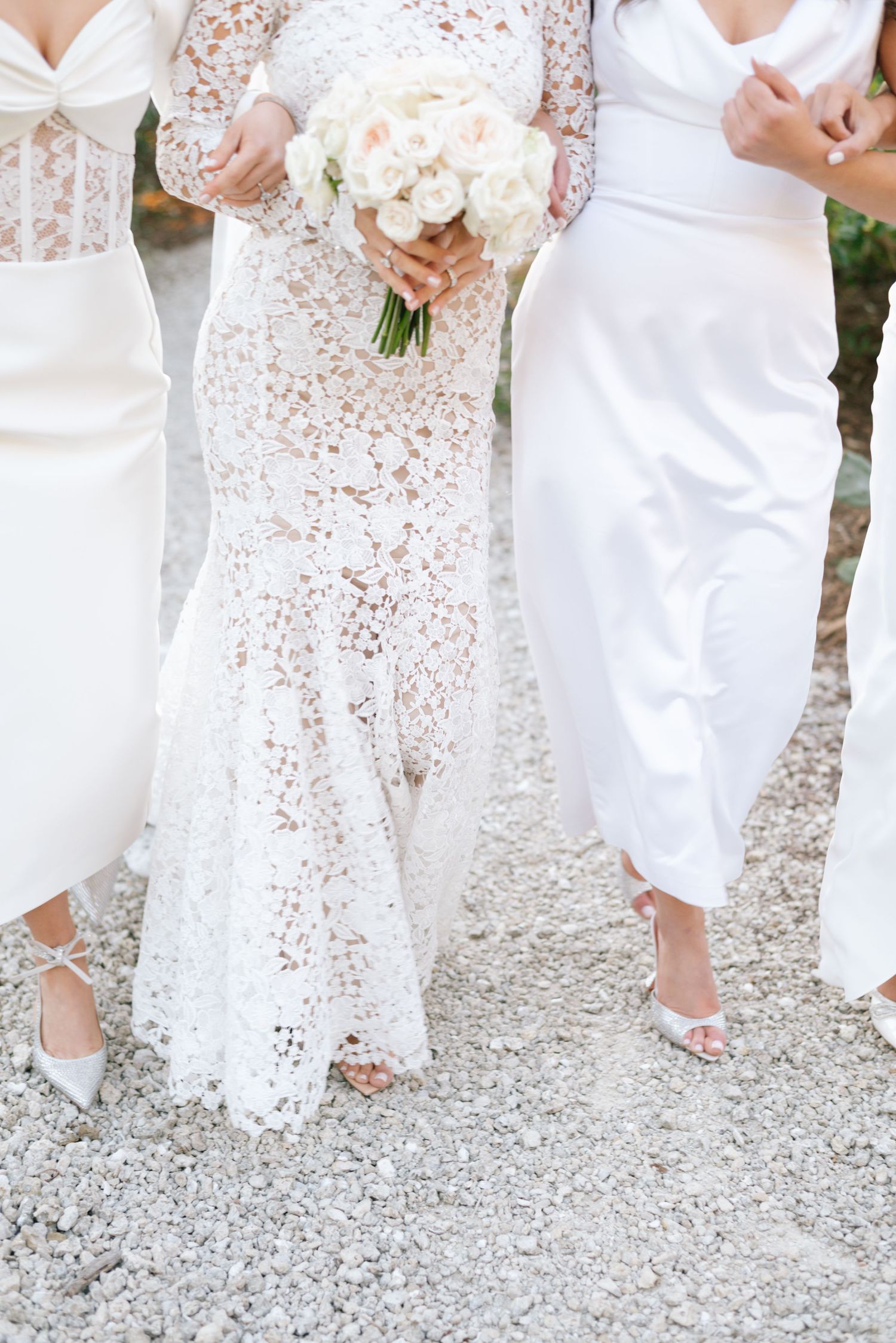 Row of bridesmaids in white lace dresses shown from waist down on gravel path.