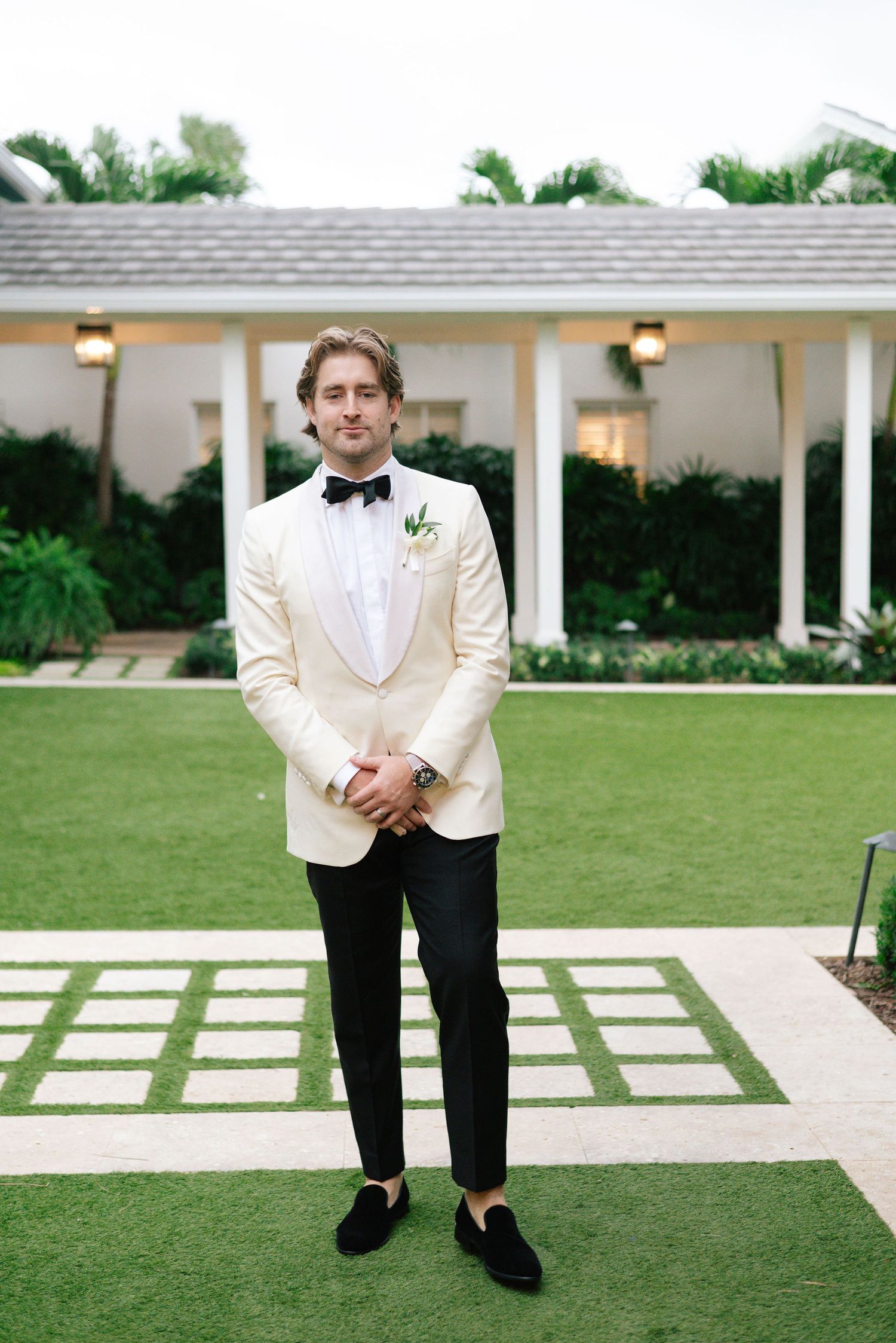 Groom in white tuxedo jacket poses on manicured lawn with colonial style architecture.