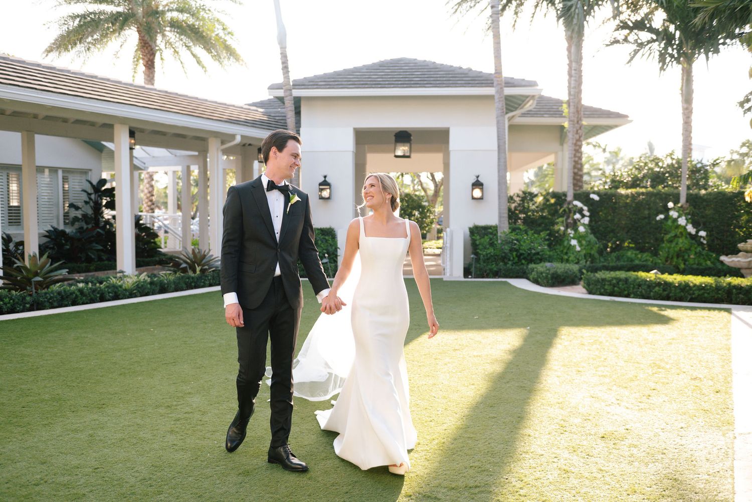 Wedding couple walking together on manicured grounds with palm trees and white architecture.