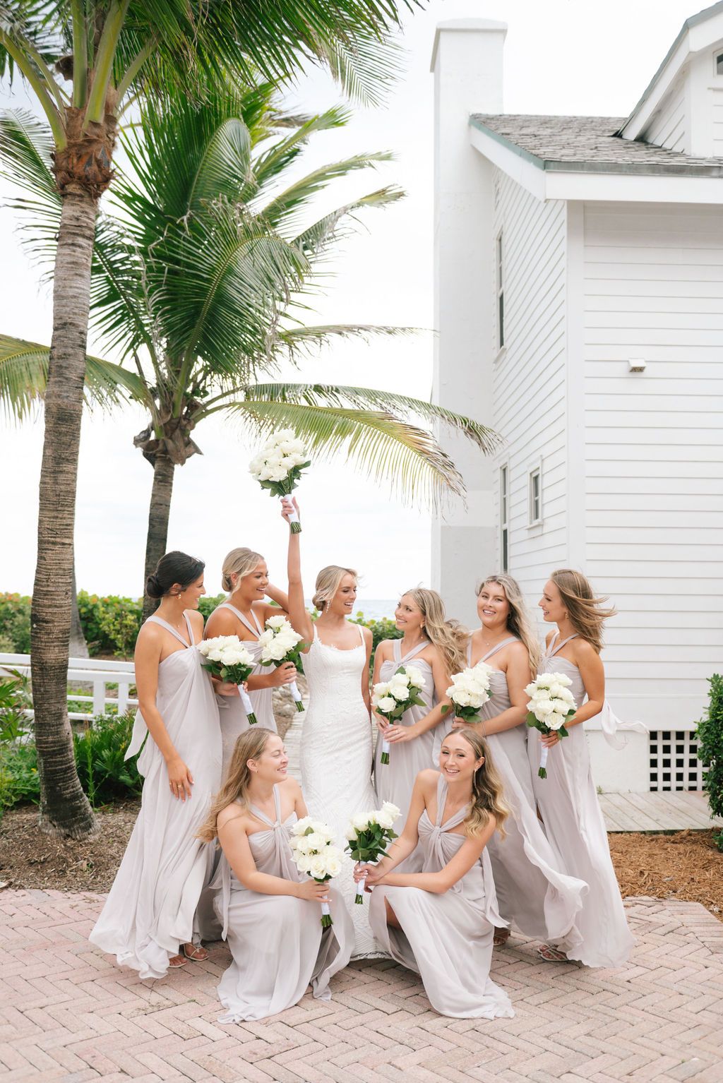 Wedding party in light gray dresses poses casually on brick pavement near palm trees and white building.