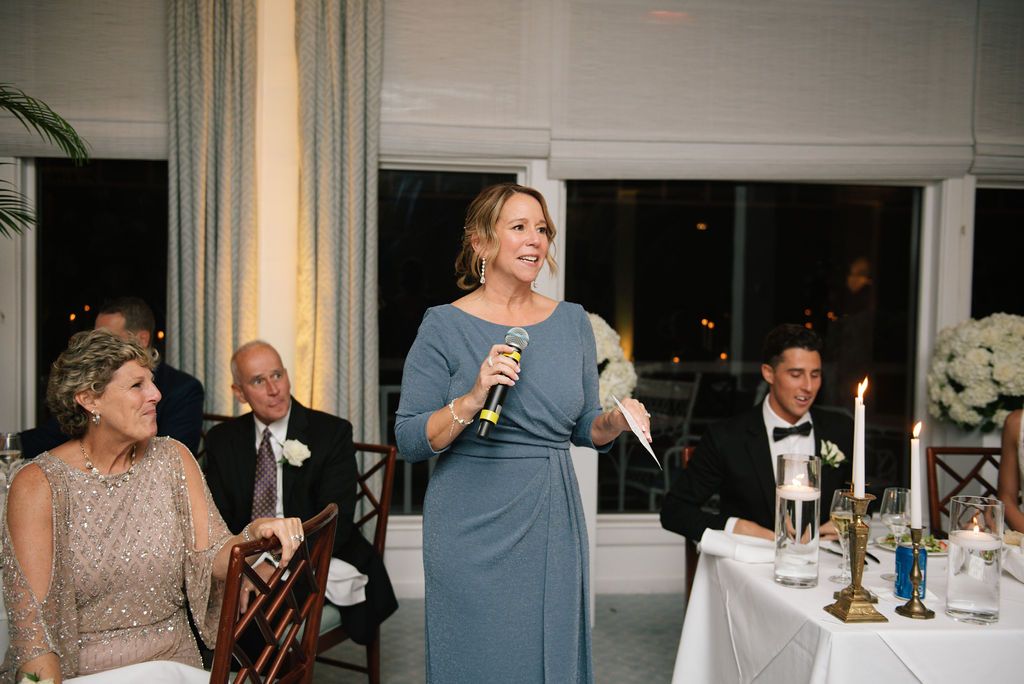 A speaker in a blue dress gives a toast during an elegant wedding reception with candlelit tables.