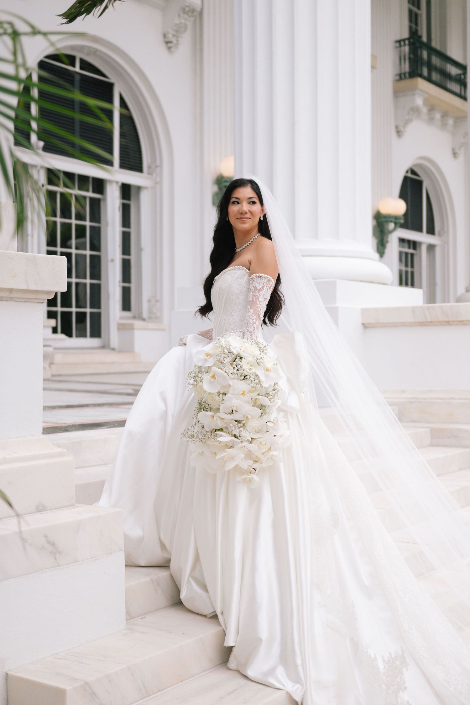 A bride in a luxurious white gown poses with her bouquet against white colonial architecture.