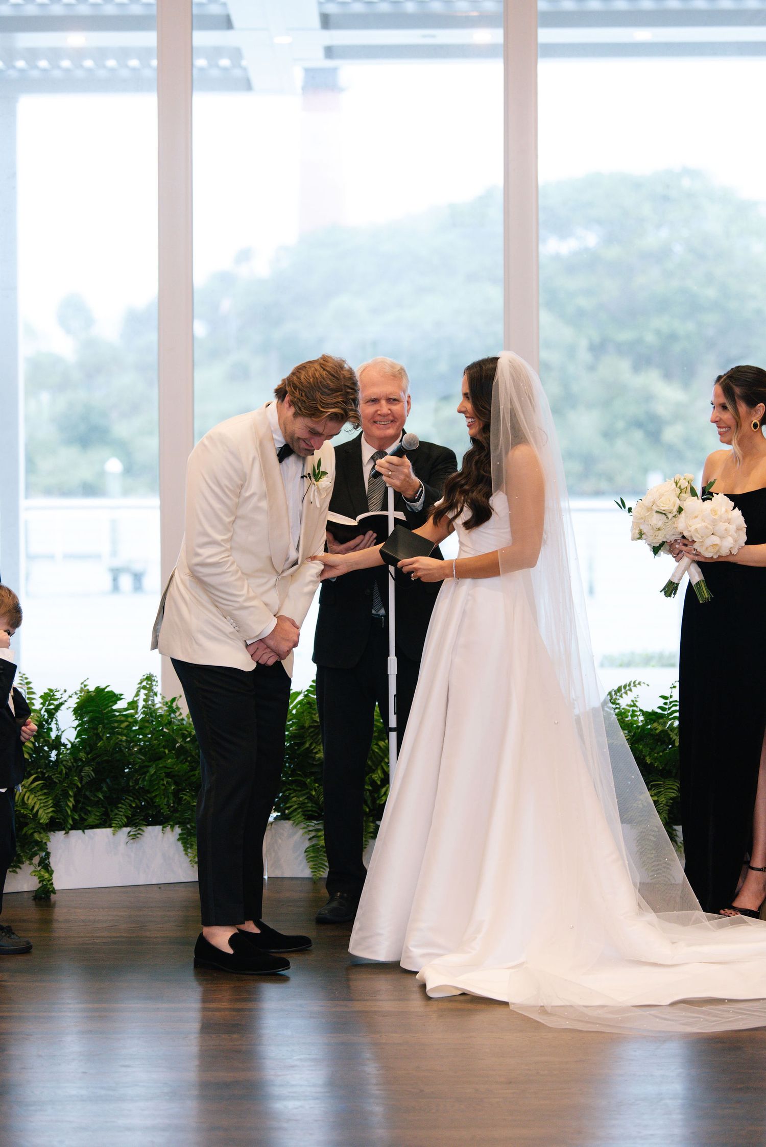 Wedding ceremony taking place in bright venue with floor-to-ceiling windows overlooking water.