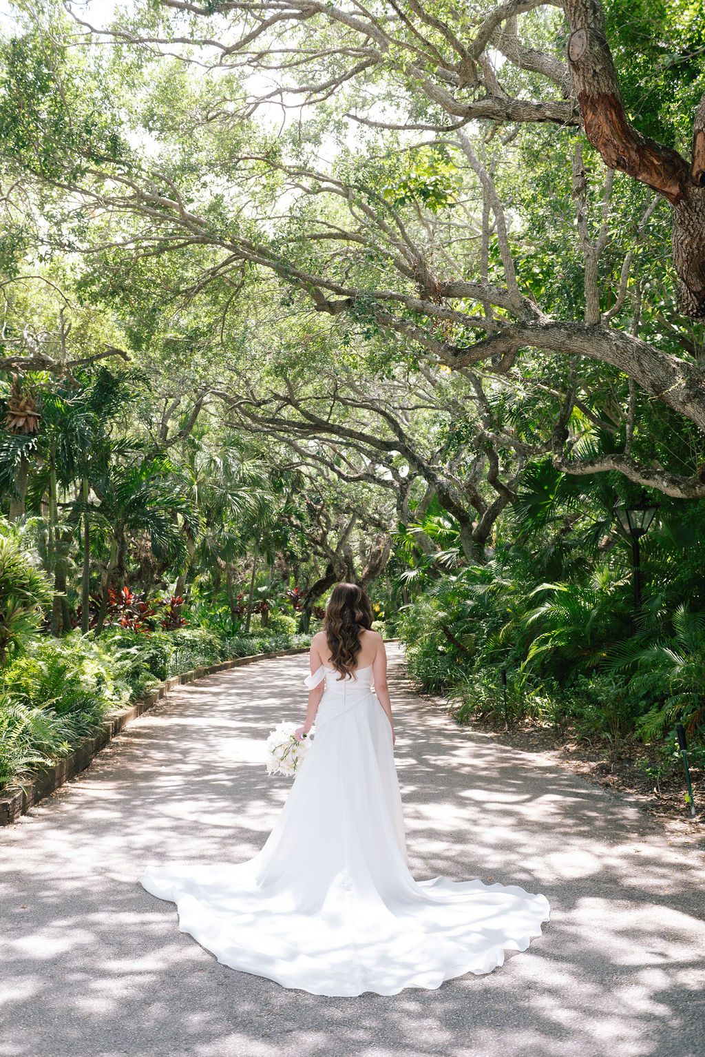 Long white wedding dress train extends down a sunlit path under tropical trees.