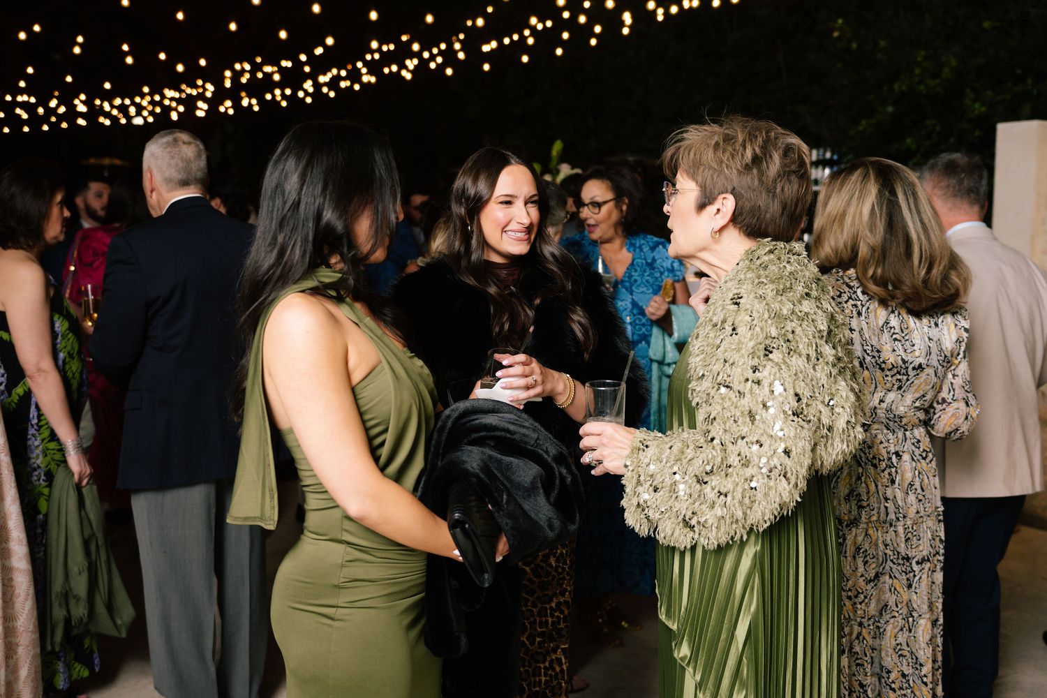Wedding guests in olive green dresses mingle at an evening reception under twinkling string lights.