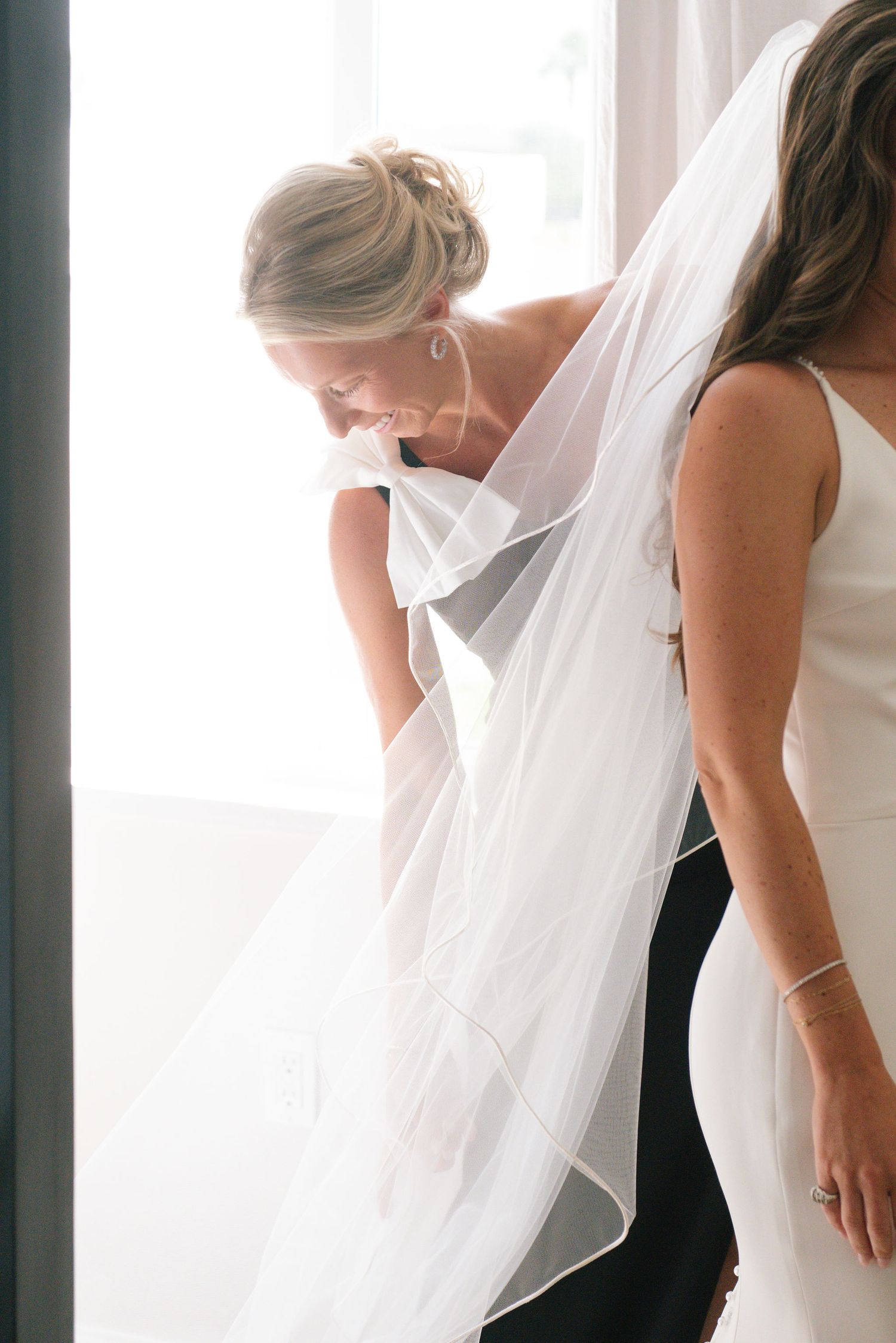A bride in a white wedding gown and veil shares a joyful candid moment with someone in black.