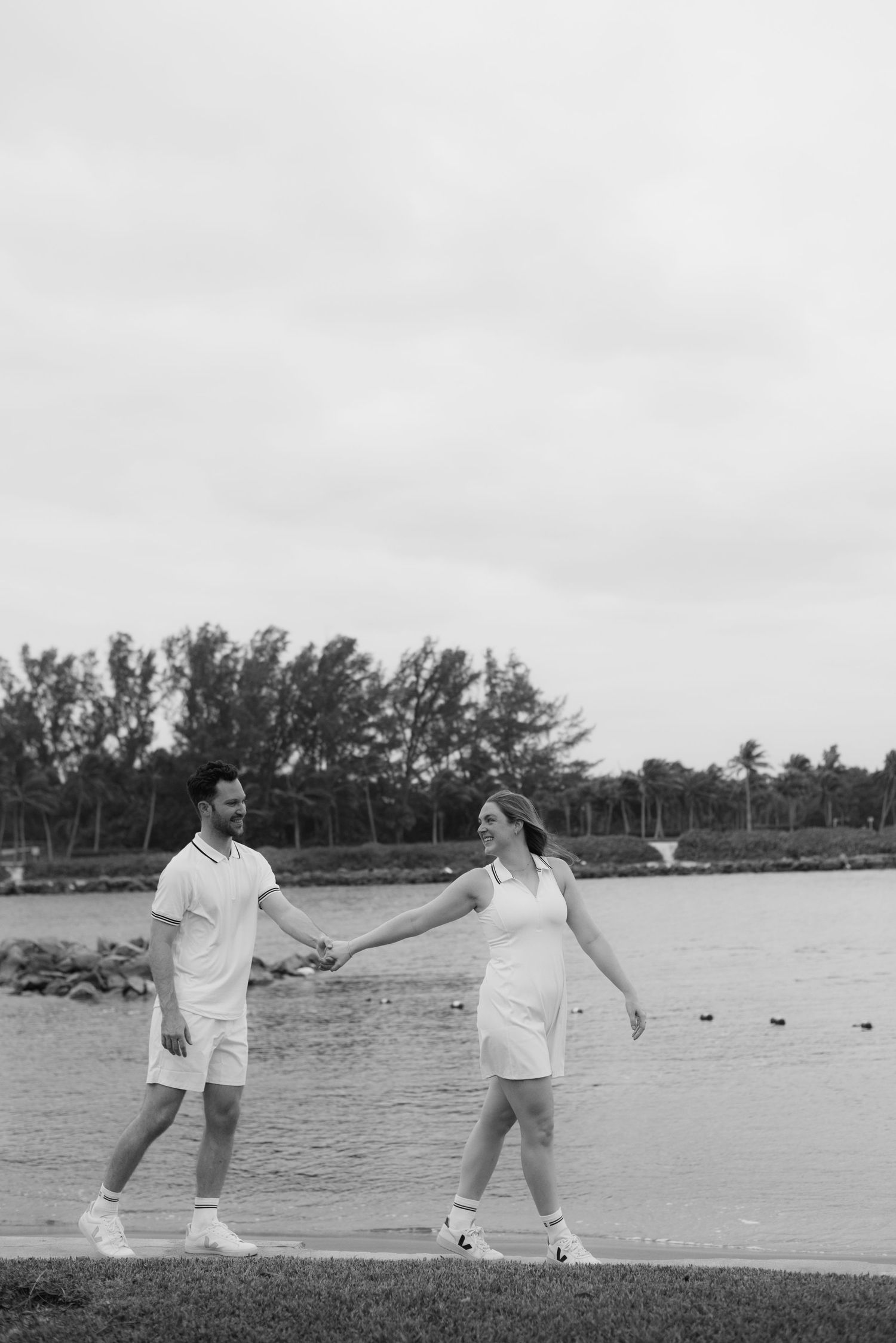 Two people in white outfits play tennis near a waterfront with trees in the background.