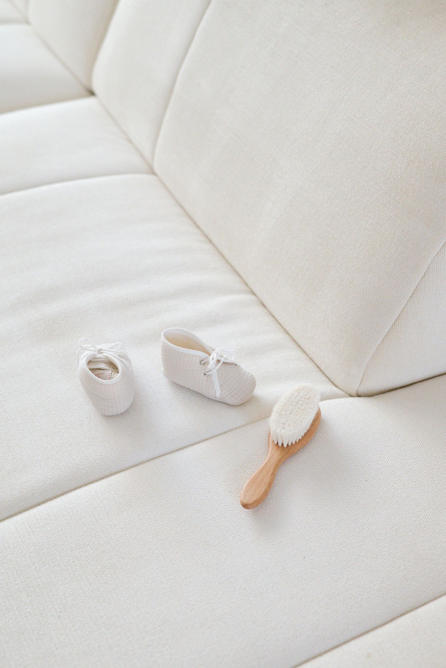 Tiny white baby shoes and wooden brush on a plush cream-colored couch.