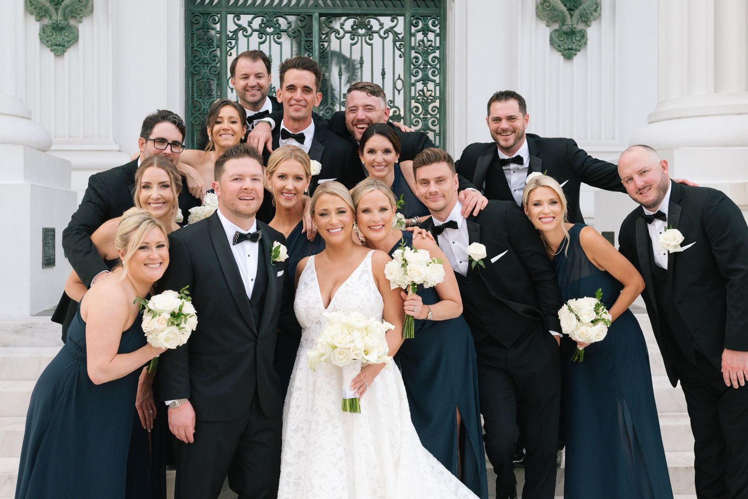 Wedding party in tuxedos and navy bridesmaid dresses poses on steps with smiling bride holding white bouquet.