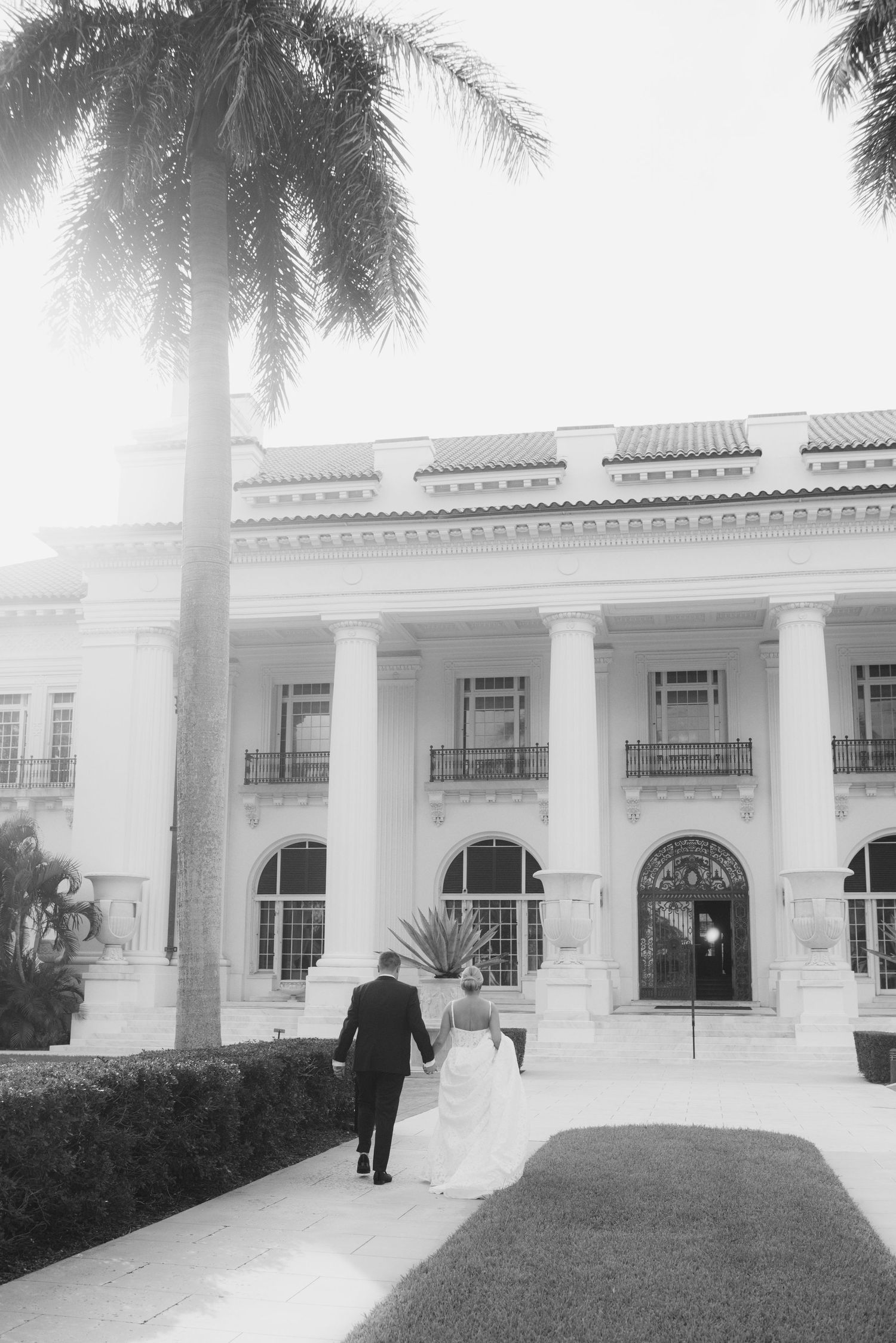 Bride and groom walking hand in hand toward an elegant columned mansion in black and white wedding photo.