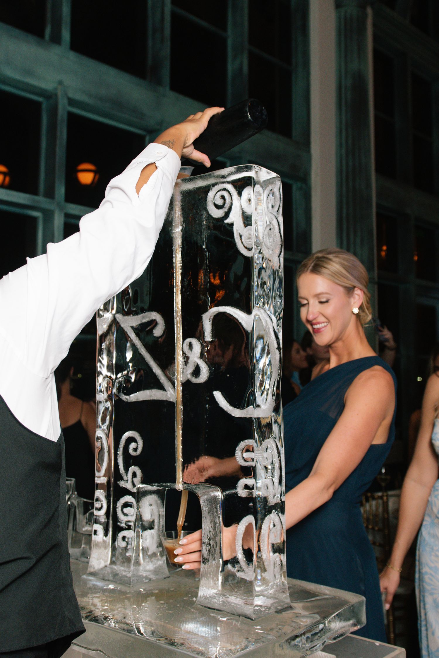 Smiling woman in navy dress enjoys a drink poured through an ornate monogrammed ice luge.