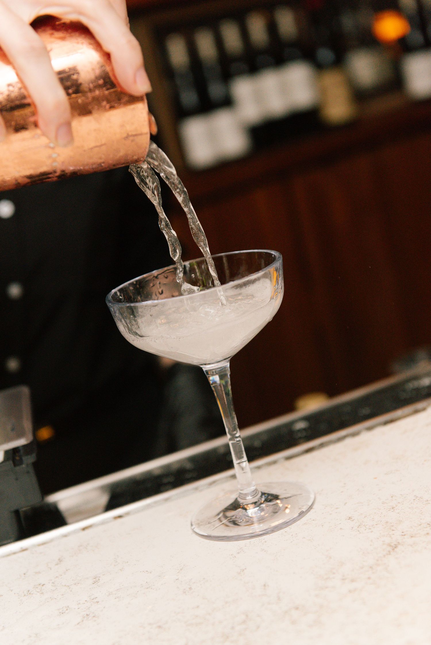 Bartender pouring a craft cocktail into a coupe glass at a marble bar counter.