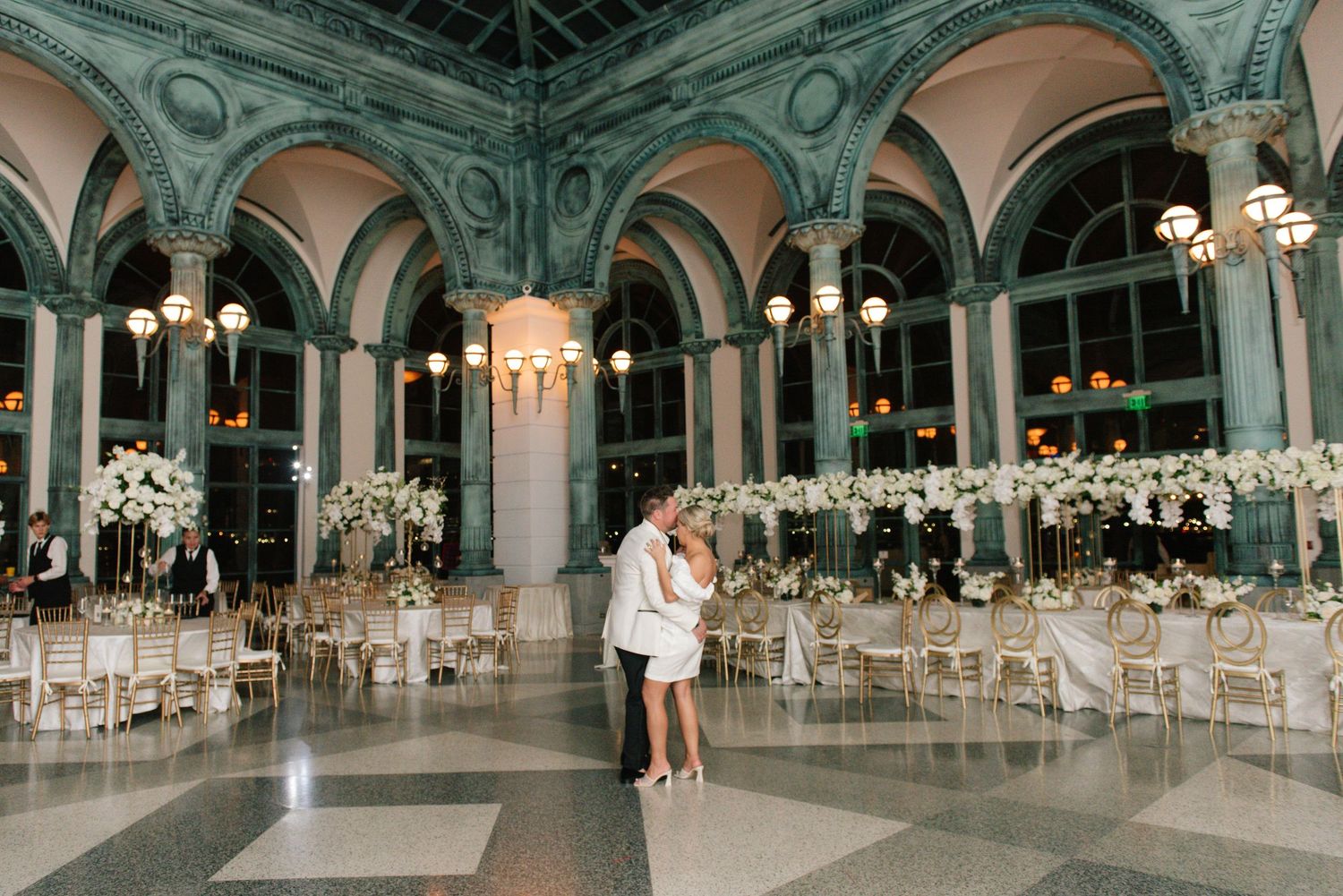 Bride and groom sharing first dance in stunning arched ballroom with floral centerpieces.